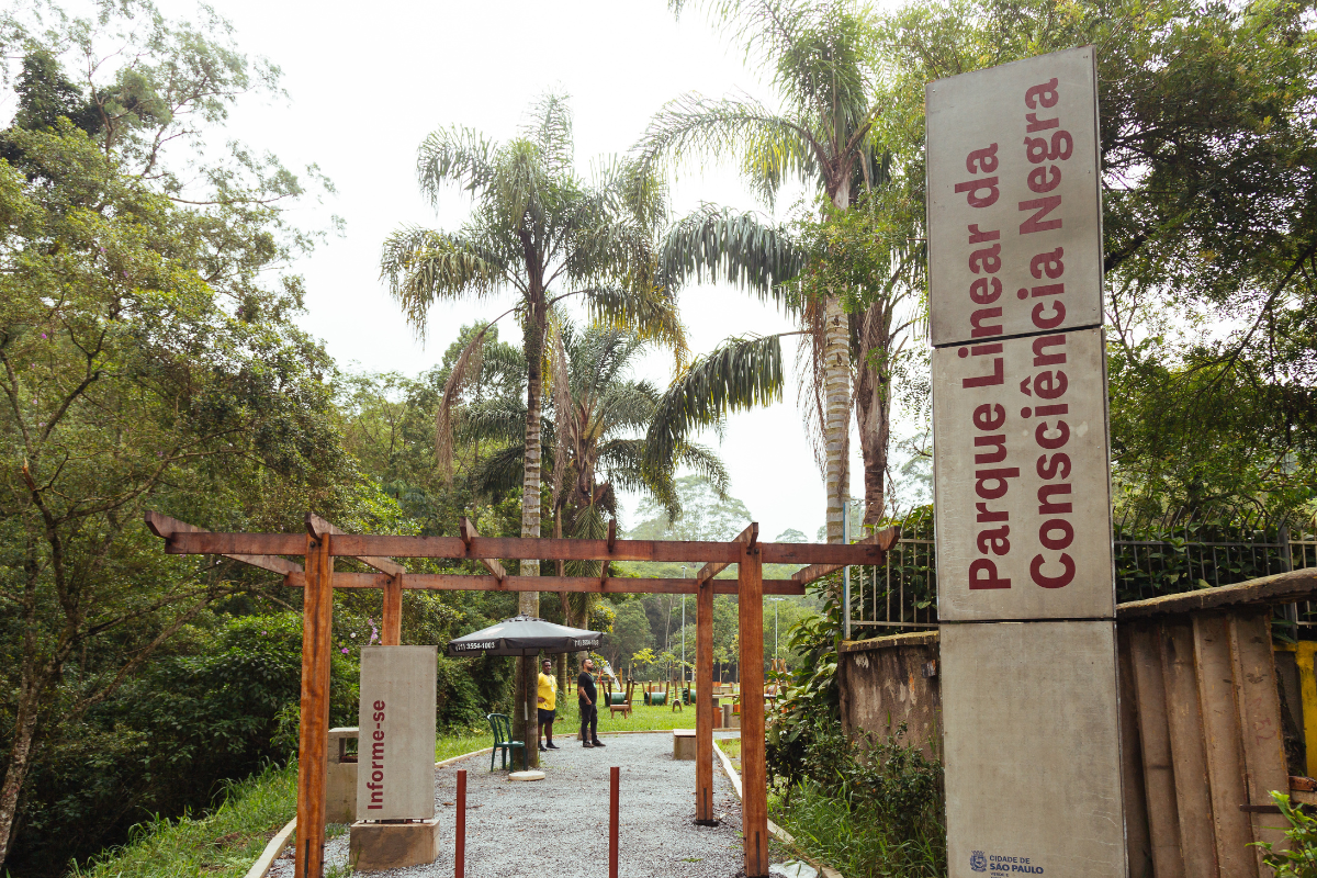 A imagem mostra a entrada do Parque Linear da Consciência Negra, localizado na cidade de São Paulo. Em destaque à direita, há um totem de concreto alto com o nome do parque escrito em letras grandes e na vertical: 
