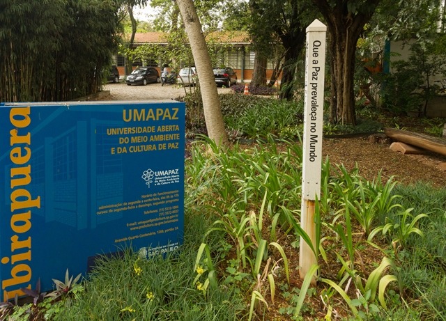 Totem com os dizeres “Que a Paz prevaleça no Mundo” instalado no jardim da sede da Universidade Aberta do Meio Ambiente e da Cultura de Paz (UMAPAZ), no Parque Ibirapuera. Ao fundo, destaque para a placa azul com detalhes em amarelo e laranja que identifica a instituição, símbolo da promoção da cultura de paz e educação ambiental em São Paulo.