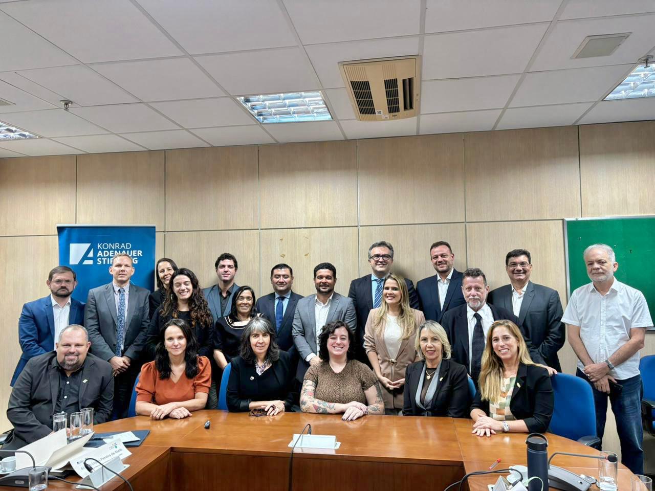 A imagem mostra um grupo de aproximadamente 22 pessoas reunidas em uma sala de reuniões corporativa. Elas estão posando para uma foto formal em frente a uma parede revestida de madeira clara, sob iluminação fluorescente. No canto esquerdo, há um banner azul com o logotipo e nome da Fundação Konrad Adenauer (Konrad Adenauer Stiftung), organização alemã de cooperação internacional. No canto direito, há um quadro verde parcialmente visível. As pessoas estão organizadas em duas fileiras: uma sentada à frente, atrás de uma mesa de madeira, e outra em pé ao fundo. Estão trajando roupas formais e semiformais, com destaque para ternos, camisas sociais e blazers em cores neutras como preto, branco, cinza, bege e azul-marinho. Há diversidade de gênero e aparência entre os presentes, com expressões em geral sorridentes e postura profissional. O ambiente sugere um evento institucional, reunião de trabalho ou encerramento de curso com foco em políticas públicas ou formação política, possivelmente ligado à atuação da fundação mencionada.