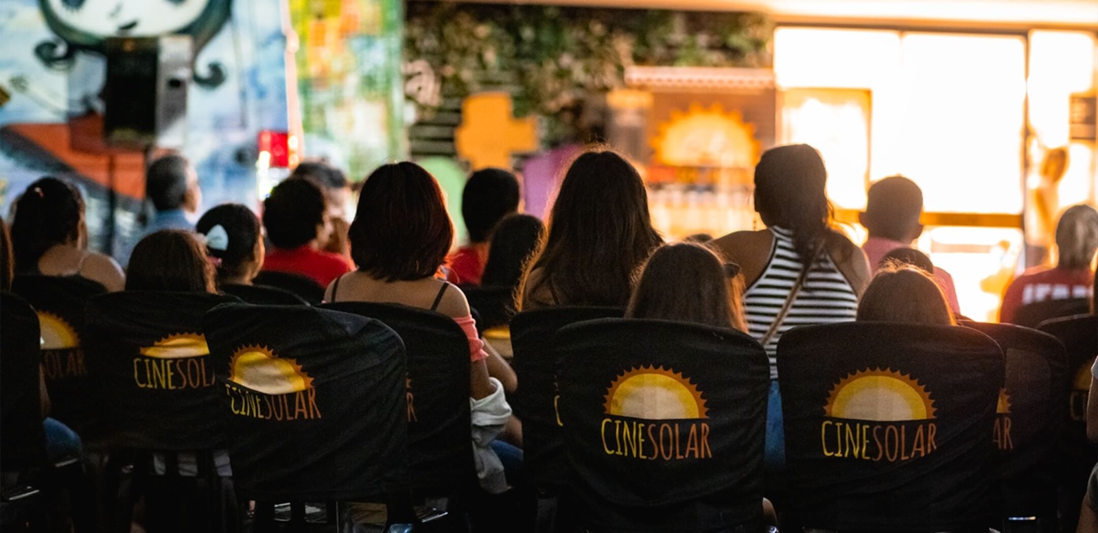 A imagem mostra uma plateia assistindo a uma sessão de cinema em um ambiente cultural decorado com grafites e luzes suaves. As pessoas estão sentadas em cadeiras pretas com encostos personalizados com o logotipo do CineSolar, que traz a imagem de um sol estilizado em amarelo e laranja acima do nome do projeto, em letras amarelas e laranjas. A cena é capturada por trás do público, destacando a variedade de pessoas presentes, incluindo adultos e crianças. A iluminação do ambiente é predominantemente quente, com tons dourados vindos da tela à frente, criando um clima acolhedor e comunitário. Ao fundo, há murais coloridos com arte urbana, reforçando a proposta cultural e inclusiva do evento. O CineSolar é um cinema itinerante que utiliza energia solar para exibir filmes, promovendo cultura e sustentabilidade em diversas regiões do Brasil.