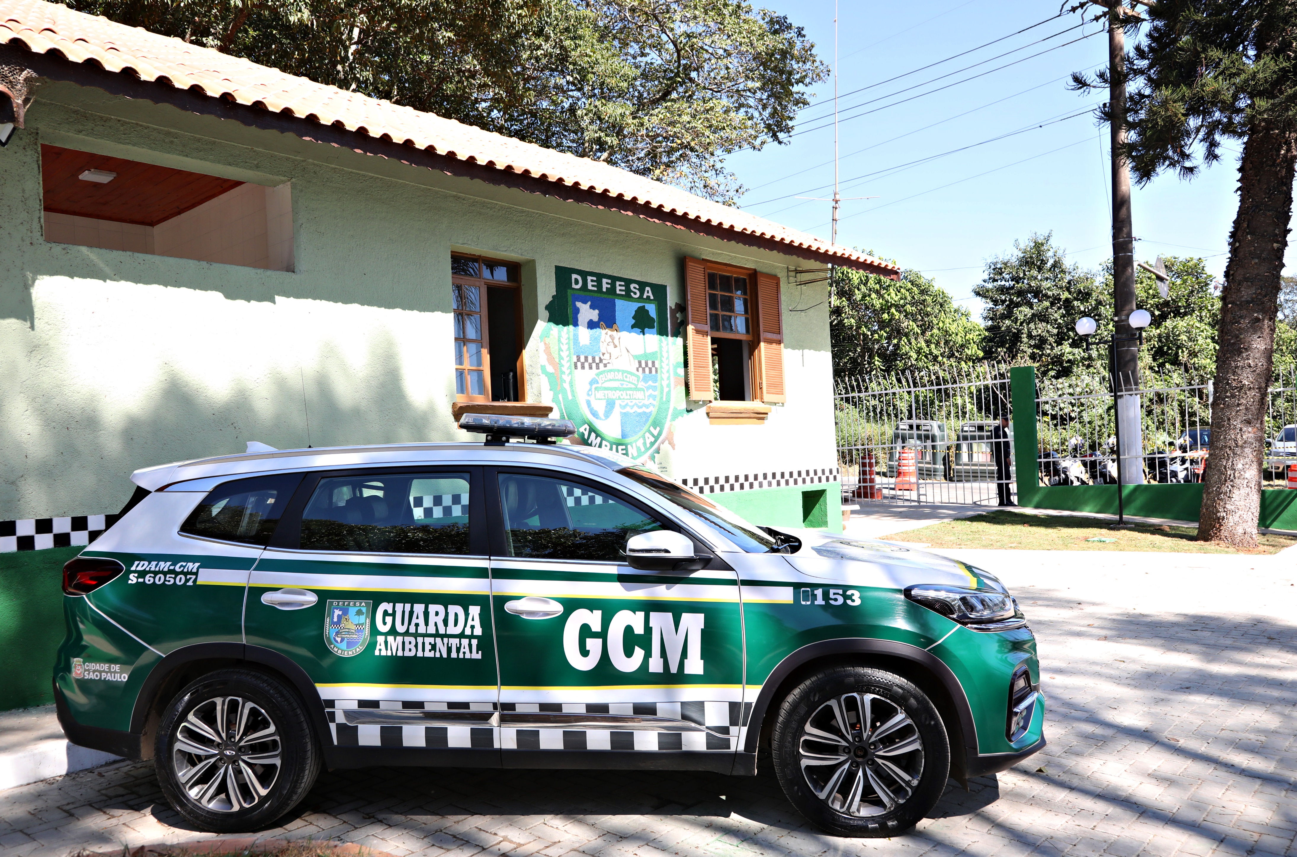 #PraTodosVerem A imagem mostra uma viatura da Guarda Civil Metropolitana (GCM) Ambiental em primeiro plano, estacionada em frente à nova Inspetoria de Defesa Ambiental da GCM. Ao fundo, é possível ver o prédio da Inspetoria, que acaba de ser inaugurada. O emblema da unidade é destacado em um escudo, com o desenho de uma onça, árvore, mapa que simbolizam a conexão com a fauna e a proteção ambiental.