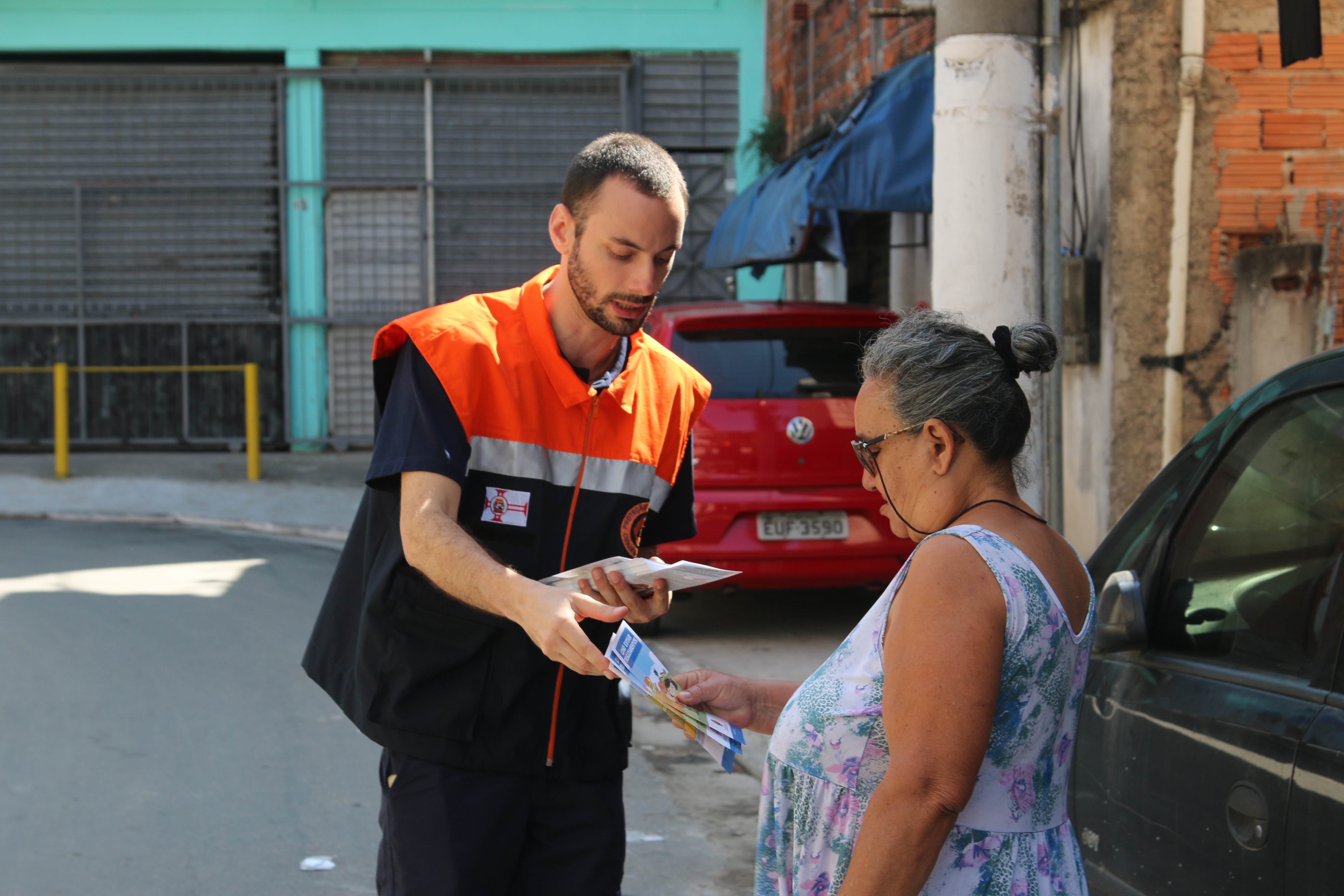 Um agente da Defesa Civil Municipal uniformizado entrega um folheto de orientações sobre áreas de risco a uma moradora da comunidade.