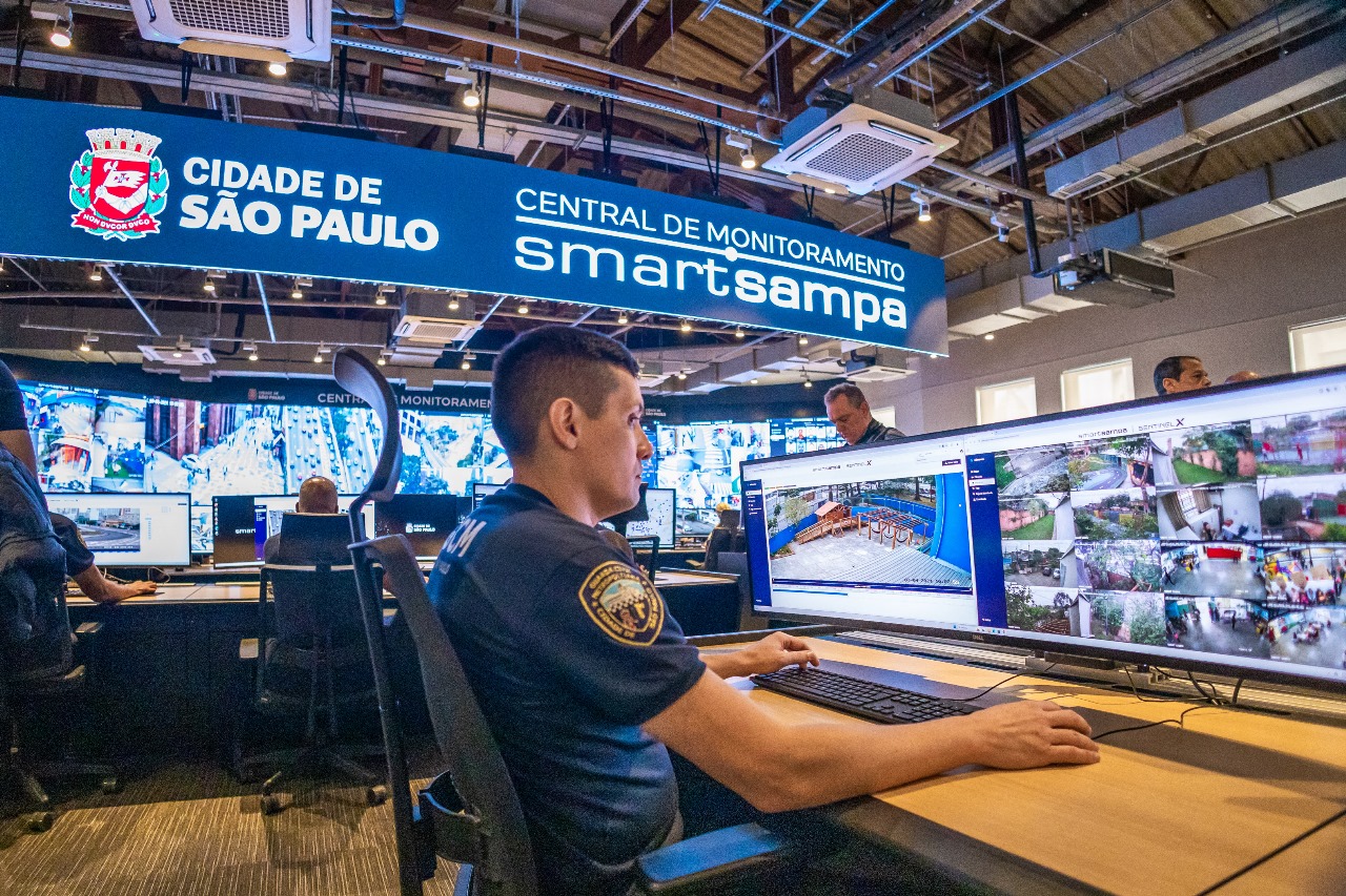 #PraTodosVerem A imagem mostra uma sala de videomonitoramento, com guardas civis metropolitanos (GCMs) sentados à frente das telas, monitorando imagens em tempo real. As telas exibem diversas imagens de câmeras de segurança, refletindo a operação em diferentes áreas da cidade. No topo da imagem, à esquerda, há um título em branco que descreve o 