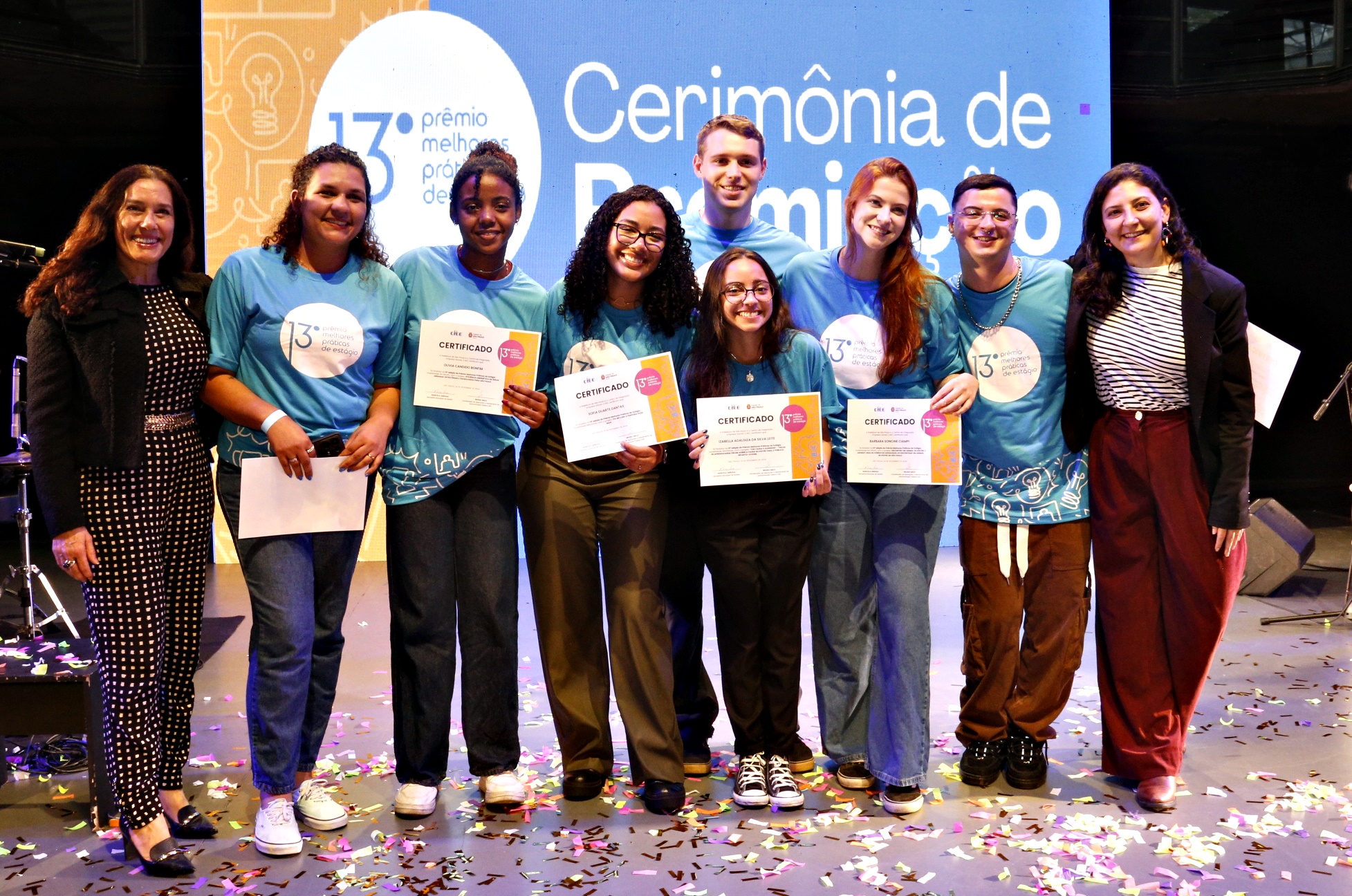 #PraTodosVerem A imagem registra seis jovens vestidos com camiseta azul, mostram seus certificados na 13ª Edição do Prêmio Melhores Práticas, acompanhados de duas mentoras, ambas com casacos pretos e cabelos soltos, em primeiro plano. Ao fundo, observa-se um painel azul com letras brancas com o nome do evento. 