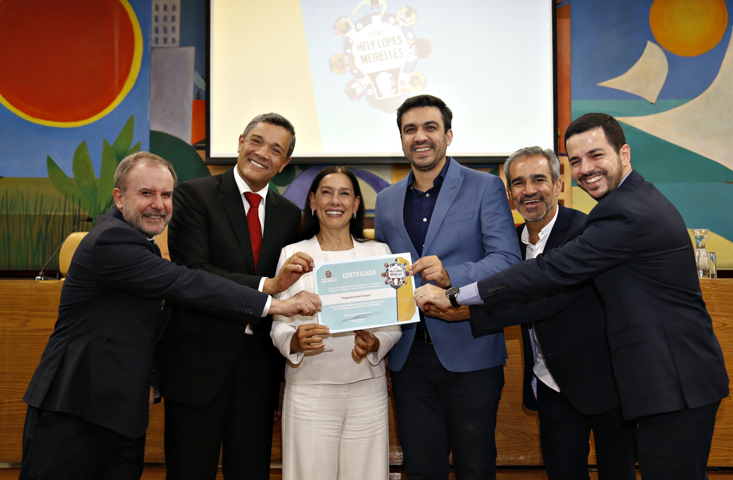 #PraTodosVerem A imagem mostra cinco homens vestidos com roupa social escura e uma mulher com roupa clara, segurando um certificado da 4ª Edição do Prêmio Hely Lopes Meirelles, em destaque. Ao fundo, o painel do evento com o logotipo do prêmio.