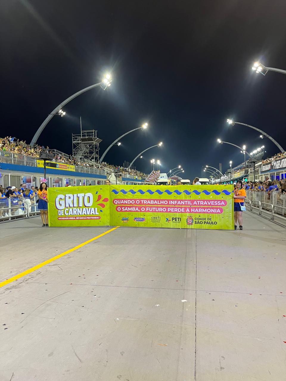 Equipe da SMADS segurando faixa do programa Proteja o Futuro. A faixa é verde e traz o slogan do Grito de Carnaval 2025: Sociedade unida, infância protegida.