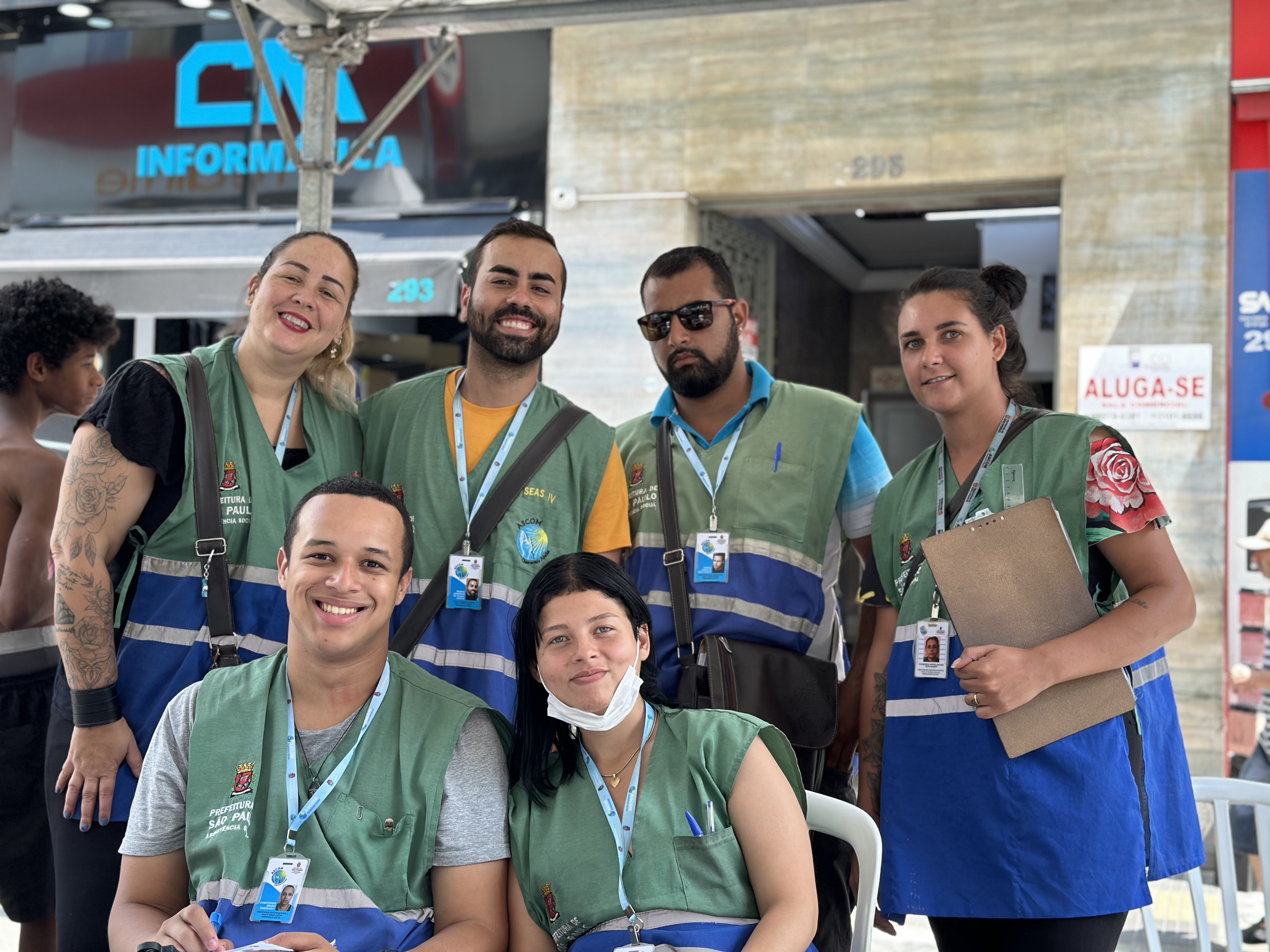 Cinco assistentes sociais, vestindo coletes verde e azul, posam sorridentes para a foto