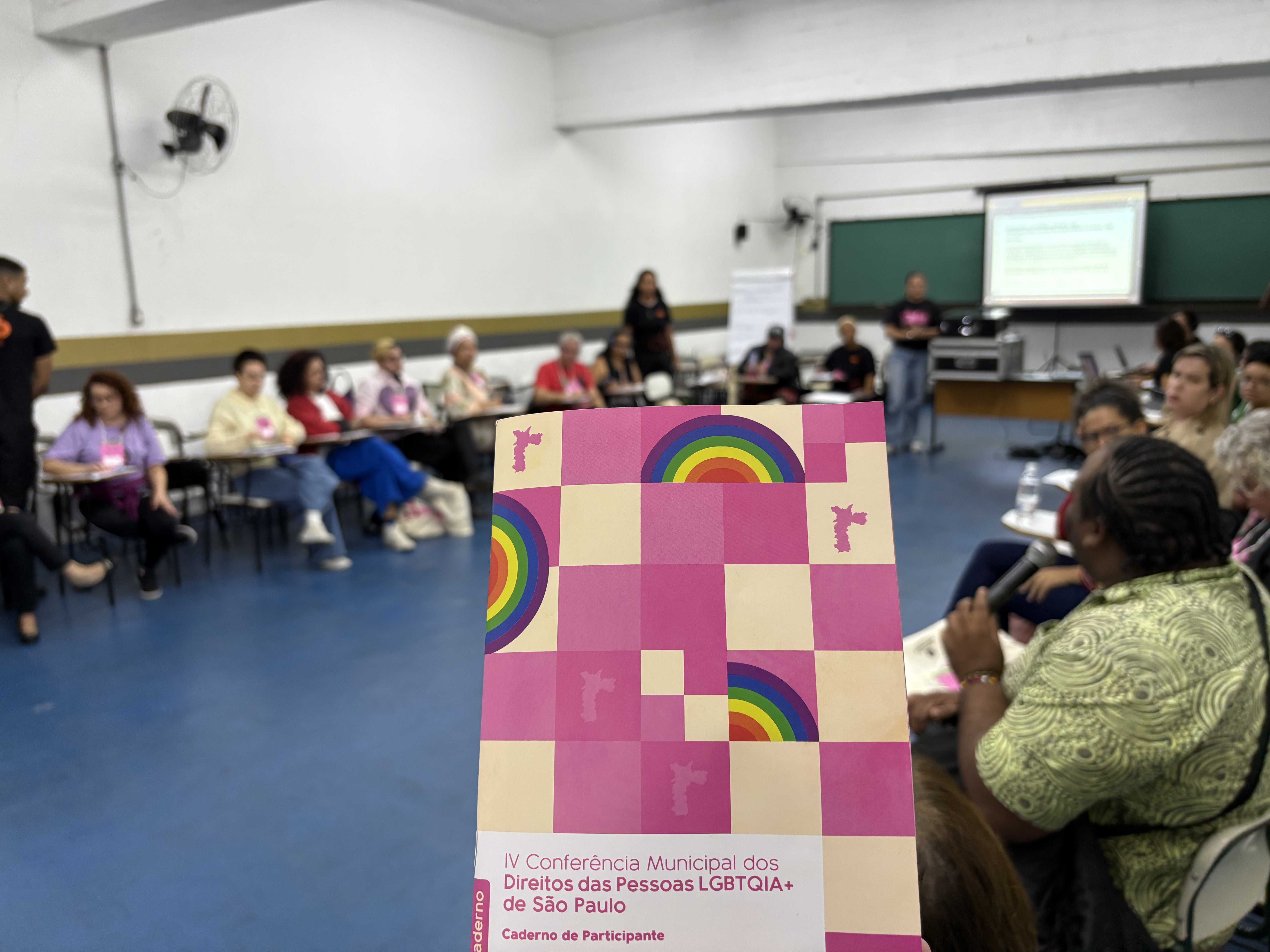 Caderno de participante da IV Conferência Municipal dos Direitos das Pessoas LGBTQIA+ de São Paulo, e ao fundo a roda de debate
