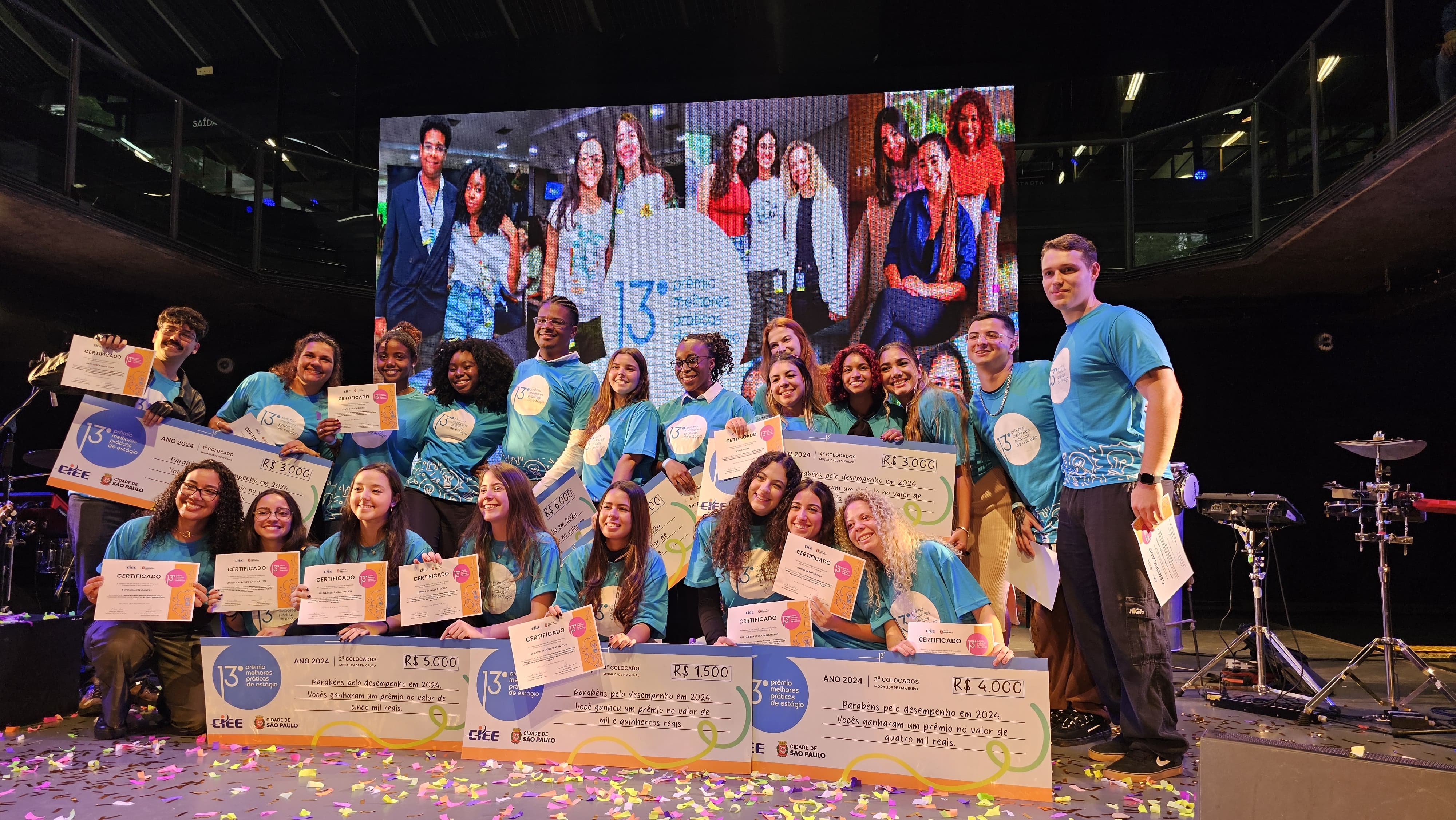 Imagem mostra os finalistas da 13ª edição do prêmio melhores práticas de estágio. De camiseta azul, eles exibem o certificado e os cheques da premiação