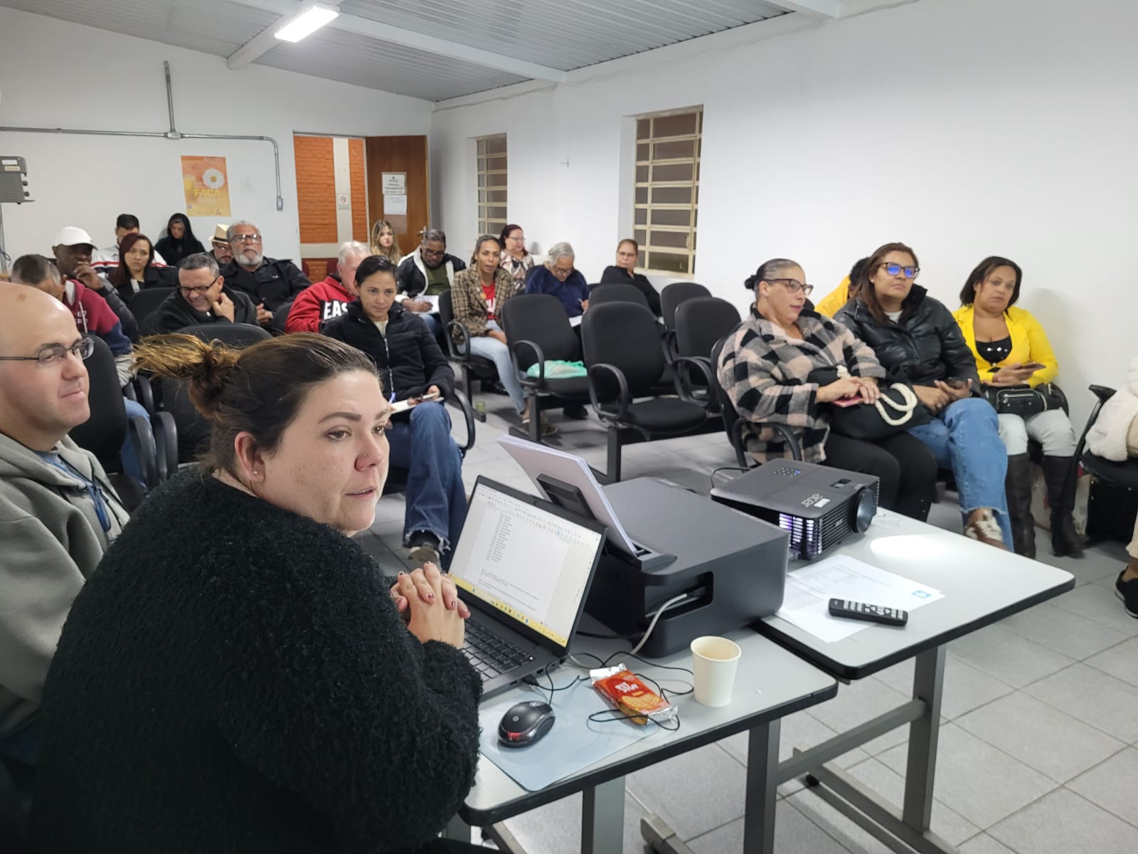 A imagem mostra uma reunião em uma sala com cerca de 25 pessoas. Em primeiro plano, à esquerda, está uma mulher de pele clara e cabelos presos, usando um suéter preto felpudo. Ela está sentada à frente de um notebook e voltada para o público, com as mãos unidas, em posição de escuta ou fala. Ao lado dela está um homem calvo, usando óculos e um moletom cinza. Sobre a mesa à frente dos dois há um projetor, um controle remoto, um copo descartável, uma embalagem de biscoitos e fios conectando os equipamentos. Ao fundo, vemos várias fileiras de cadeiras ocupadas por homens e mulheres de diferentes idades, etnias e estilos, vestidos com roupas de frio. Algumas pessoas seguram papéis ou cadernos, outras estão atentas, algumas conversando entre si ou olhando para os lados. No canto direito, três mulheres estão sentadas lado a lado, com destaque para uma de casaco quadriculado, outra de jaqueta preta e a terceira de blazer amarelo vivo. A iluminação da sala é clara e o ambiente parece ser institucional, com paredes brancas e janelas com grades. A atmosfera sugere uma reunião comunitária ou uma atividade formativa.