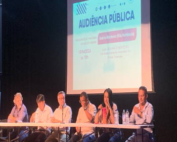 Seis Componentes na mesa falam na Audiência Pública. Um grande painel logo atrás expõe tópicos sobre a regularização fundiária