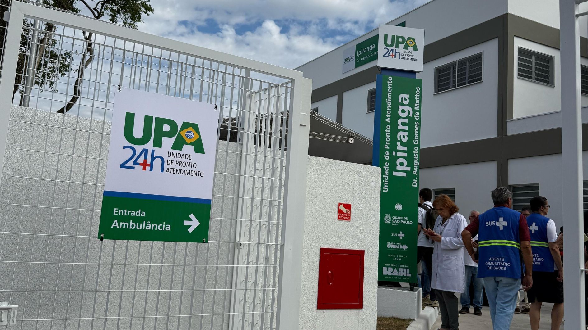 Imagem da entrada da Upa, com uma placa dizendo: UPA 24h. Unidade de Pronto Atendimento. E no fundo alguns funcionários do SUS