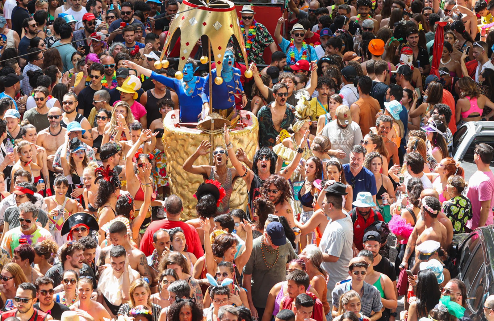 Cena de um bloco carnavalesco na avenida. Pessoas fantasiadas e pulando e acenando