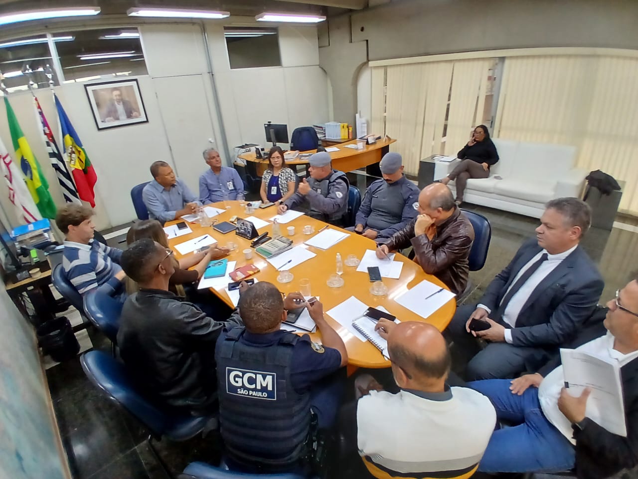 A imagem mostra uma sala de reuniões com uma grande mesa oval de madeira no centro, rodeada por cadeiras. Há cerca de 15 pessoas sentadas à mesa, a maioria vestindo roupas formais de negócios. Algumas pessoas parecem estar em uniformes, possivelmente policiais ou de segurança. Há algumas pastas e papéis sobre a mesa, e parece que uma reunião está em andamento. No fundo da sala, há um sofá branco e uma mulher sentada nele, parecendo observar a reunião. Há também bandeiras na parede, sugerindo que pode ser uma reunião governamental ou oficial. A iluminação na sala é brilhante, e a atmosfera parece formal e séria. A imagem é tirada de um ângulo ligeiramente elevado, dando uma visão geral da sala de reuniões e dos participantes.