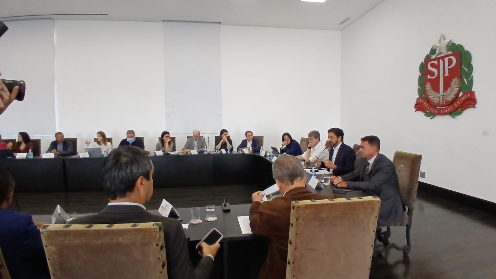 A imagem mostra uma reunião em uma sala de conferências. Uma longa mesa de conferências está no centro da sala com pessoas sentadas em ambos os lados. Há um brasão do governo do estado de São Paulo na parede atrás deles. A sala parece formal e profissional.