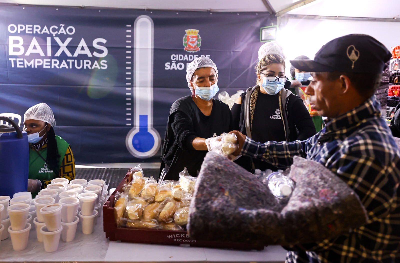A imagem mostra uma bancada com copos lacrados e pães recheados dentro de uma cesta. Acima possui uma mulher entregando dois pães recheados para um rapaz de boné segurando um cobertor.