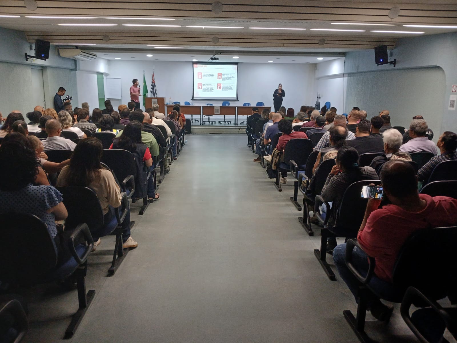 A imagem mostra um auditório e um corredor central e ambos os lados com pessoas de costas sentadas olhando para uma tela com apresentação de dados da audiência. Ao fundo da imagem, do lado direito, tem duas bandeiras e a tela na parede, com um homem de camisa rosa falando ao microfone e, do lado oposto, uma mulher de roupa preta fazendo sinais de LIBRAS.