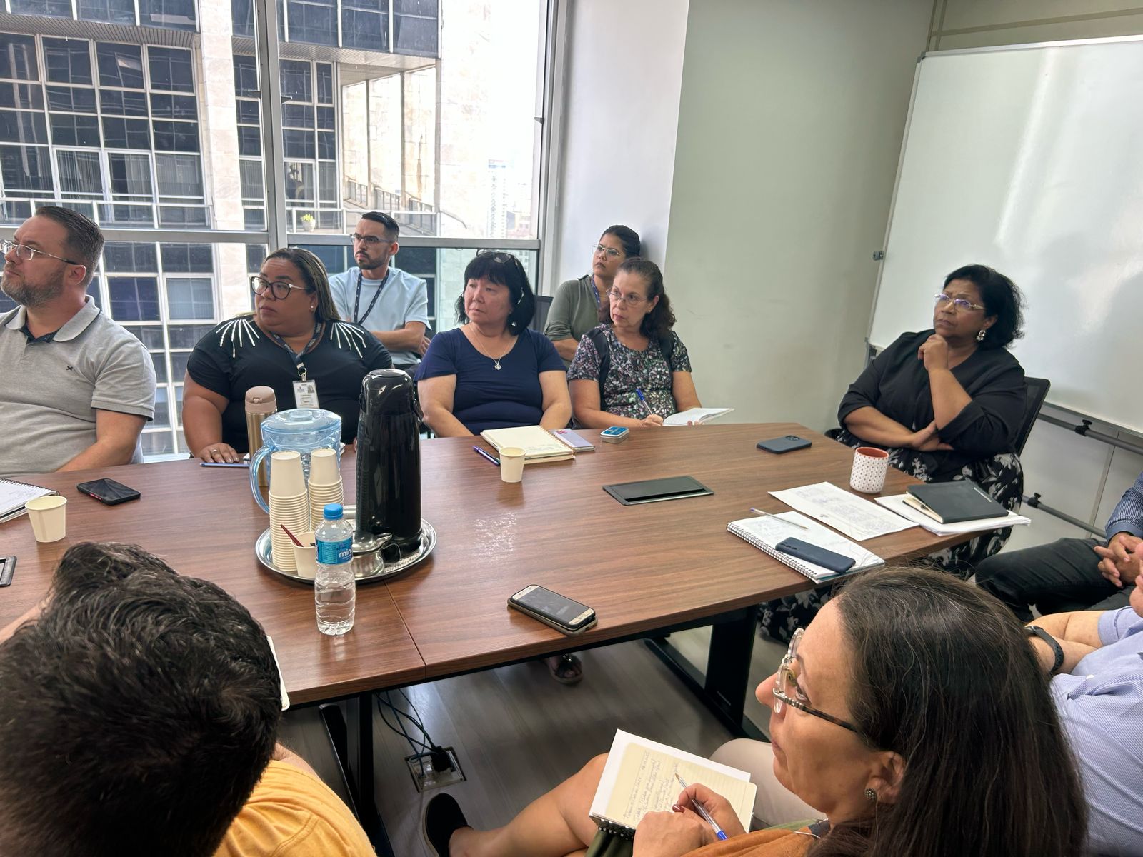 A imagem mostra um grupo de pessoas participando de uma reunião em uma sala corporativa. Elas estão sentadas ao redor de uma mesa com cadernos, celulares, papéis e uma caneca. Algumas pessoas estão fazendo anotações, enquanto outras escutam atentamente. O ambiente tem um quadro branco ao fundo e janelas que mostram prédios comerciais, sugerindo um escritório em uma área urbana. 