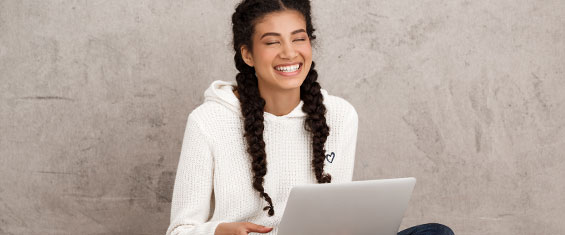 Jovem negra com cabelos trançados, vestindo tricot branco e sorrindo em frente a um computador.