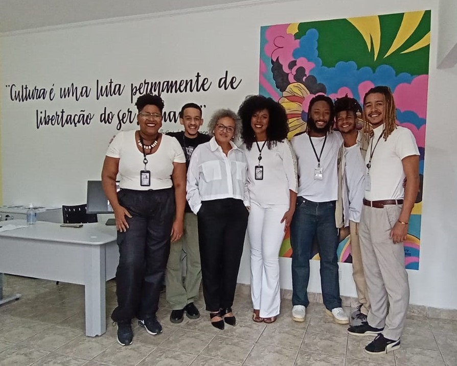 A imagem mostra a coordenadora do CRPIR São Miguel Paulista, com outras 6 pessoas da equipe, na recepção do equipamento, em frente a uma parece grafitada com imagem colorida e frase de Abdias do Nascimento 