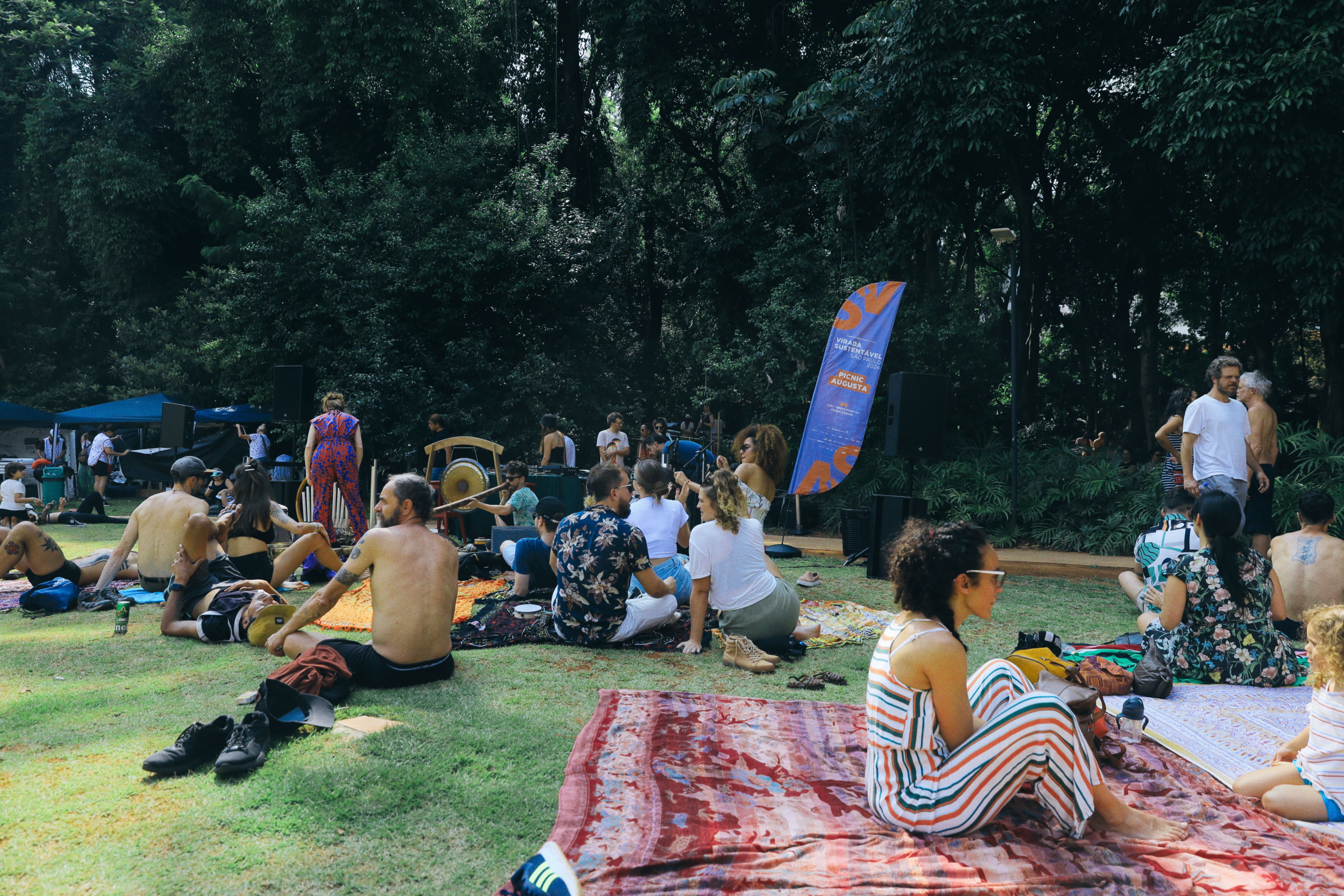 Pessoas sentadas sobre toalhas em grama, curtindo músicas e um dia ensolarado, no Parque Augusta.