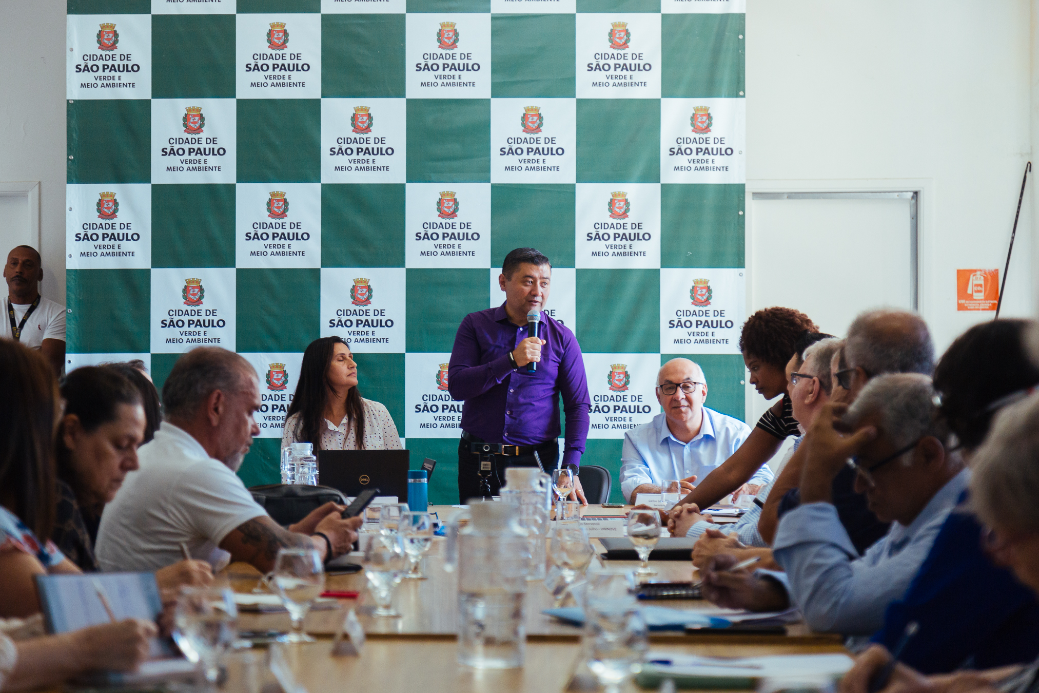 Imagem do secretário Rodrigo Aschiushi em destaque, segurando um microfone durante reunião do CADES. Ele está em frente de uma mesa de madeira, onde há diversas pessoas sentadas em cadeiras à frente da mesa, assistindo o secretário palestrar. 