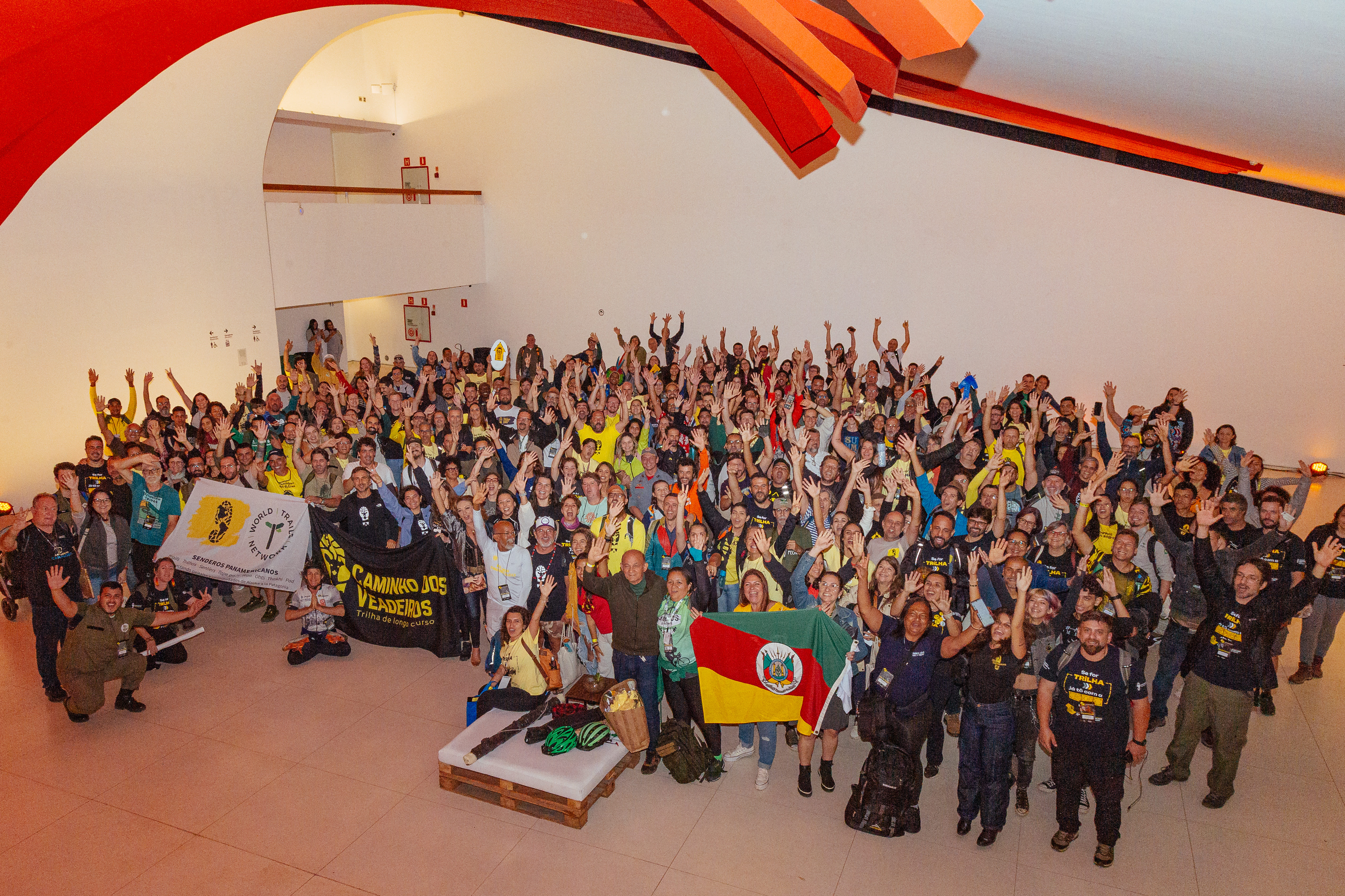 Grupo de pessoas reunidas em um salão, segurando bandeiras, e com as mãos levantadas para o alto. Elas comemoram juntas o mesmo evento chamado Congresso Brasileiro de Trilhas.  