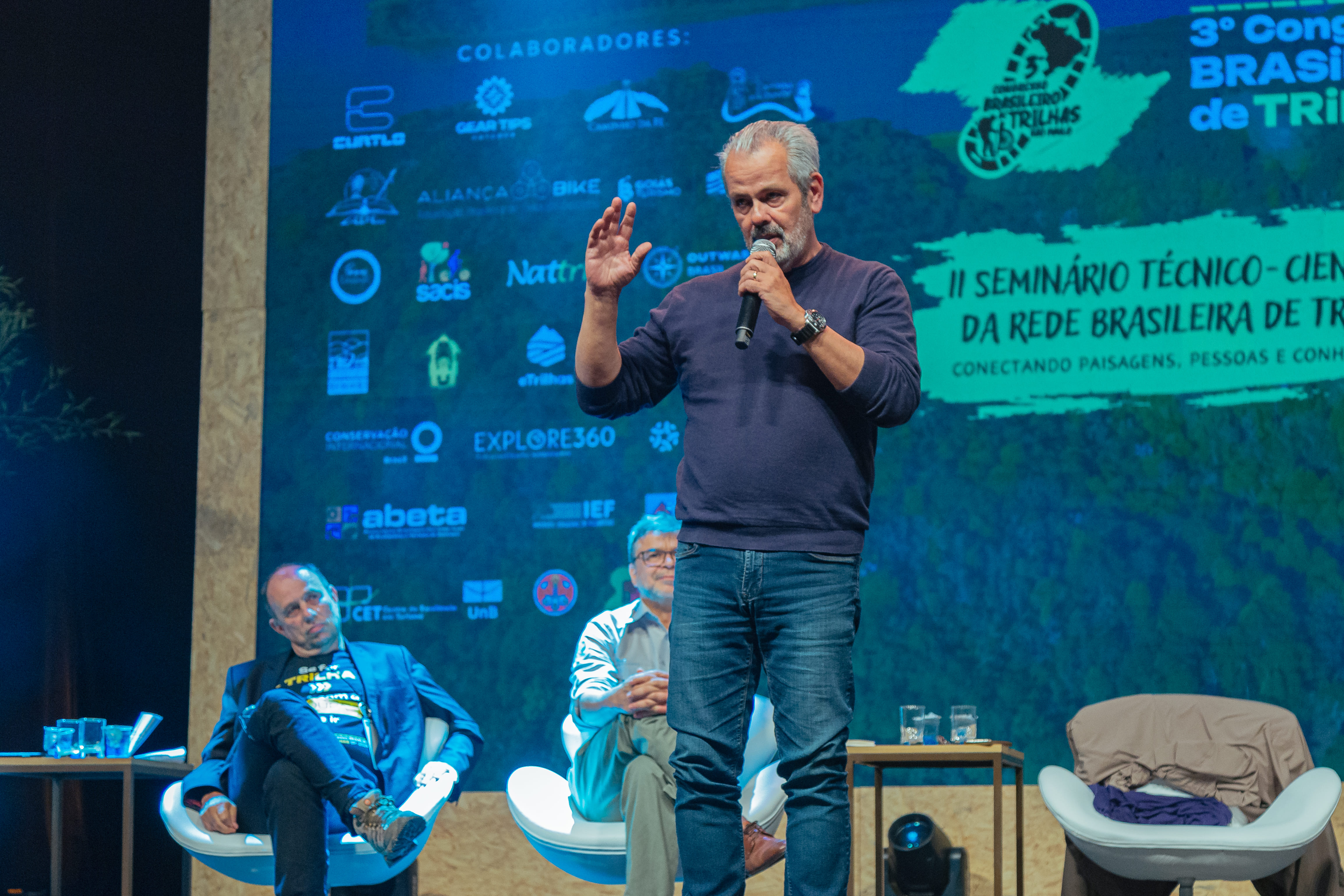 Secretário Rodrigo Ravena segurando microfone e olhando para o público, enquanto discursa durante o 3 Congresso de Trilhas. Atrás dele, há dois homens sentados em cadeiras, vestidos de roupas sociais. Os três personagen são figuras importantes. 