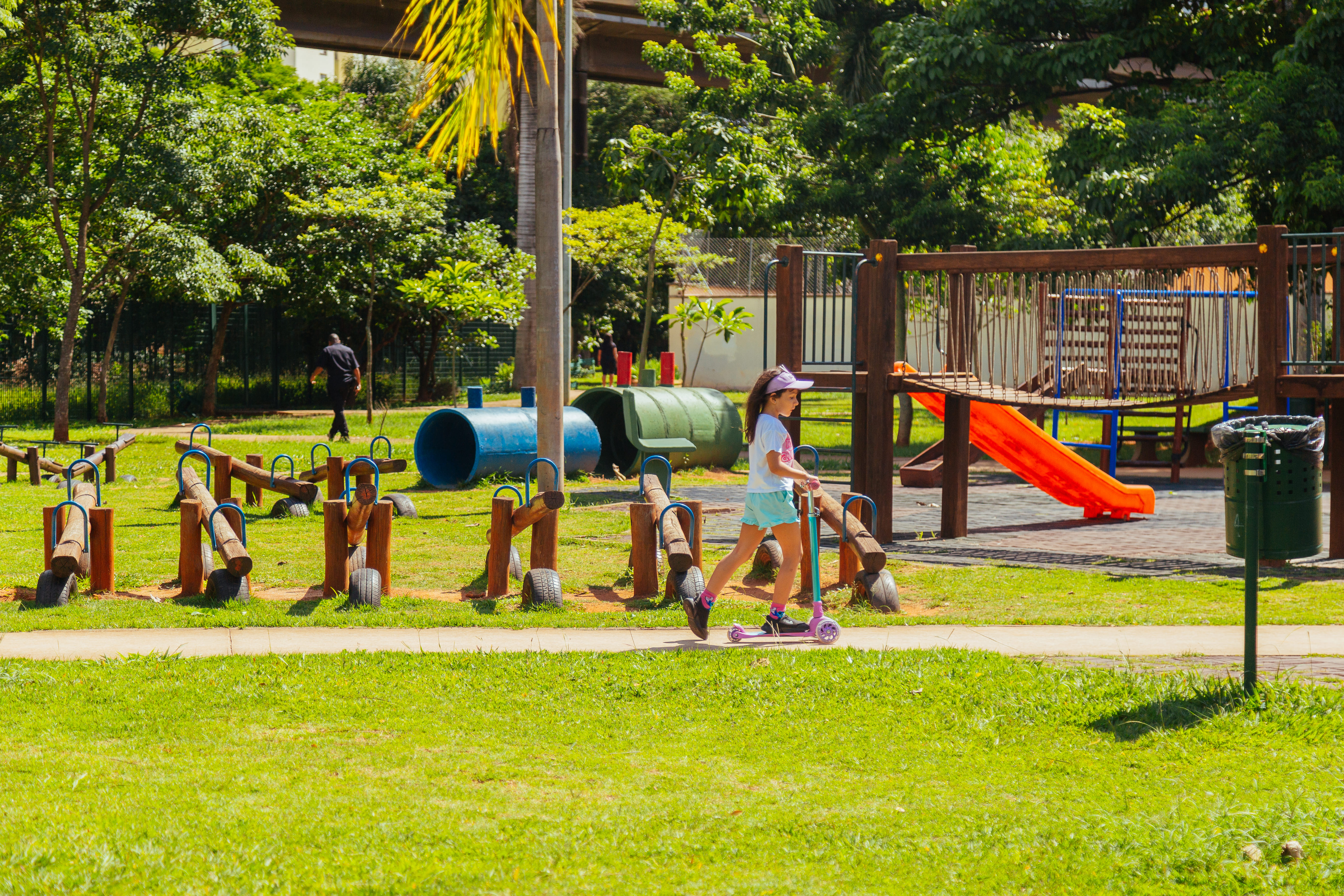 Área verde com brinquedos em parquinho de criança, com árvores ao fundo. Em destaque, uma criança anda de patins em pista do parque, próximo aos brinquedos.  