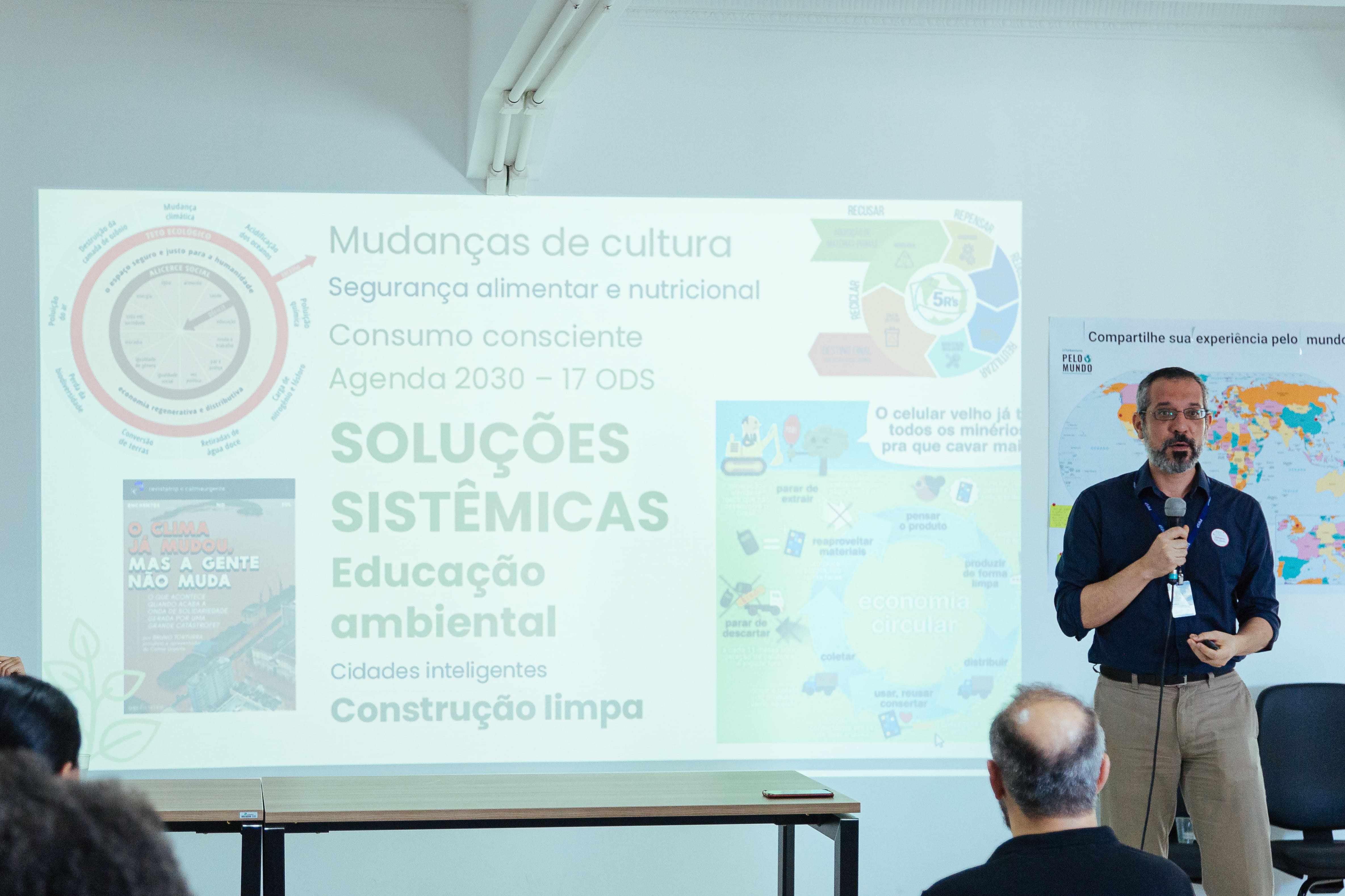 Homem ao lado de telão com apresentação sobre o Plano Municipal de Educação Ambiental. Ele segura o microfone enquanto palestra. 