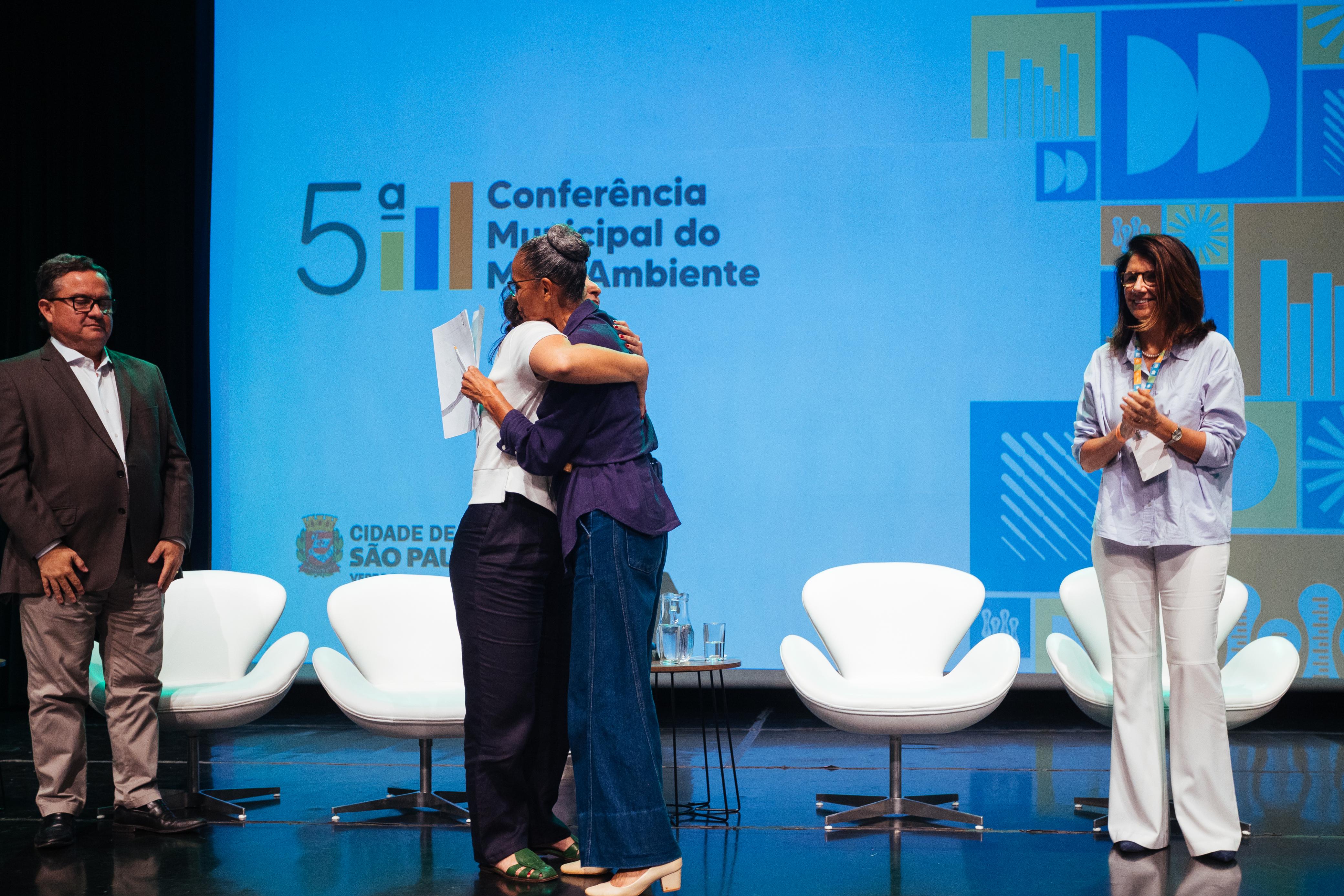 Tamires Oliveira, secretária do Verde e do Meio Ambiente de São Paulo, abraçando a ministra do Meio Ambiente, Marina Silva, durante evento. Ao fundo, uma tela de apresentação com os seguintes dizeres "5° Conferência Municipal do Meio Ambiente". 