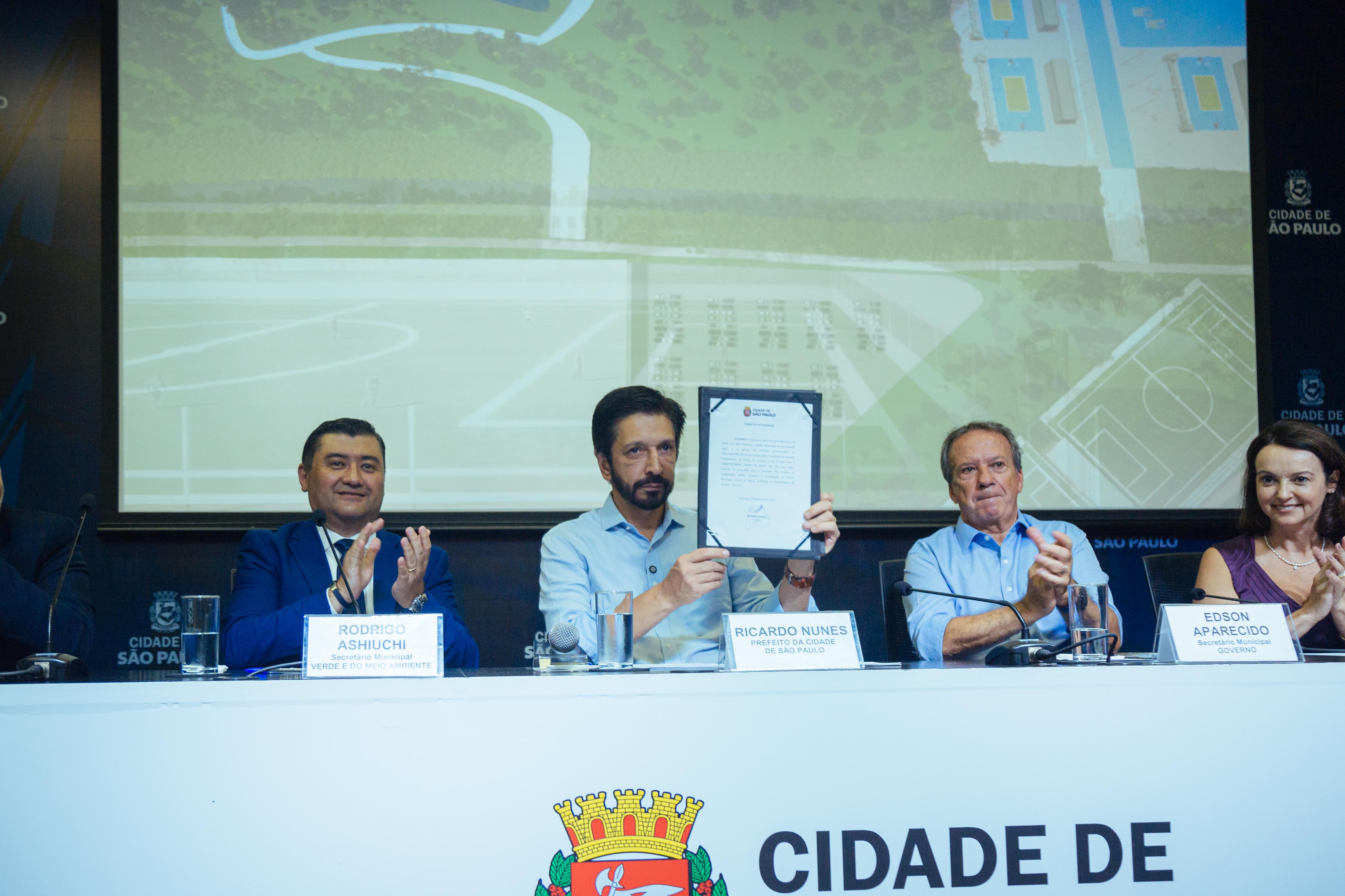 Secretário do Verde e do Meio Ambiente, Rodrigo Ashiuchi; Prefeito da Cidade de São Paulo, Ricardo Nunes; e Edson Aparecido, Secretário Municipal do Governo, sentados em frente a uma mesa, anunciando a concessão do Parque Campo de Marte. O prefeito, Ricardo Nunes, está no meio da mesa, em destaque, segurando um contrato.    