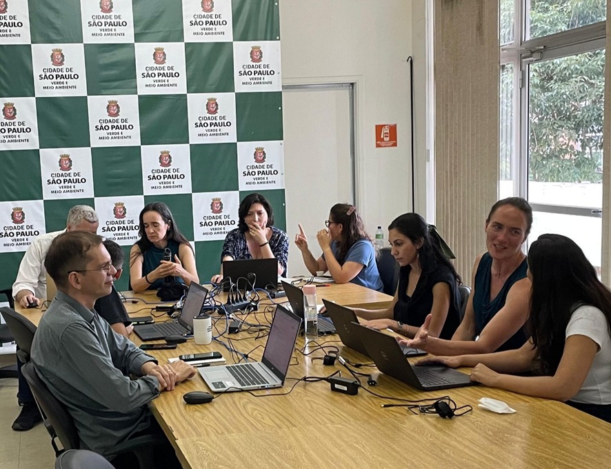 Equipe da Secretaria do Verde e do Meio Ambiente (SVMA) durante reunião de planejamento na sede da pasta. Ao fundo, painel institucional com o brasão da cidade de São Paulo e os dizeres “Cidade de São Paulo – Verde e Meio Ambiente”, em fundo verde e branco. A cena mostra a troca colaborativa entre os(as) servidores(as), com destaque para os notebooks conectados sobre a mesa.