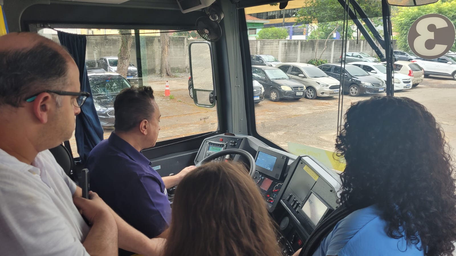 Imagem de pessoas dentro de um ônibus, de costas. Um homem está sentado no banco de motorista enquanto encara o volante do ônibus. 