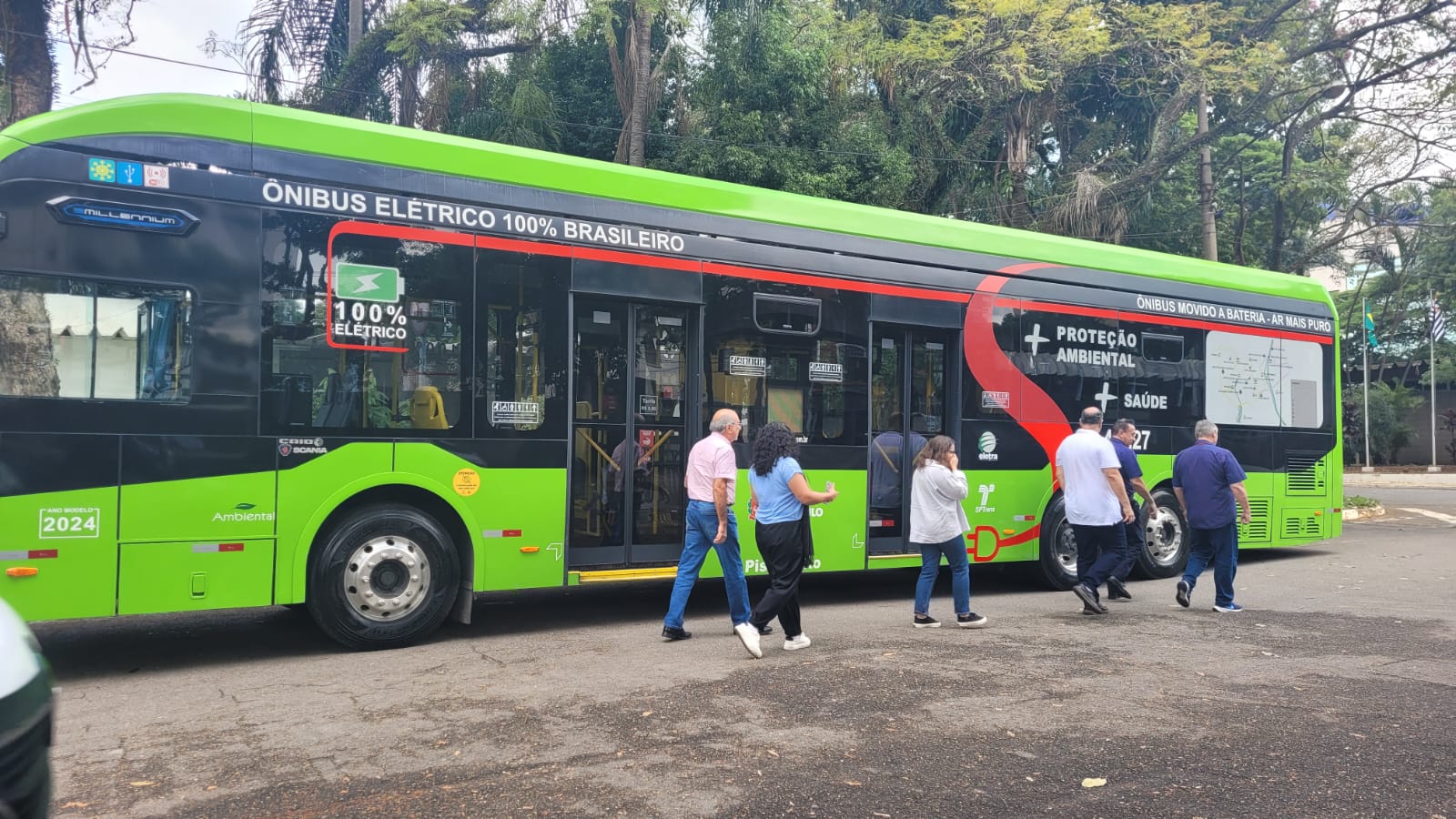 Imagem de ônibus elétrico de cor verde estacionado com pessoas andando em direção a ele. Ao fundo, várias árvores de cor verde escuro. 