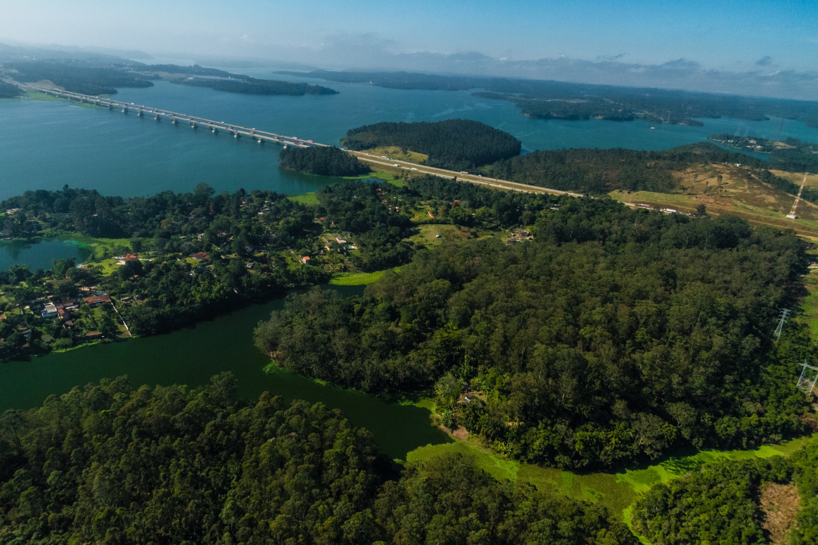 Vista aérea da Represa Billings, na zona sul de São Paulo, com destaque para áreas de mata nativa, ocupações residenciais e a ponte da Rodovia dos Imigrantes ao fundo. A região abriga importantes fragmentos de Mata Atlântica e é fundamental para o abastecimento de água e a regulação climática da cidade.