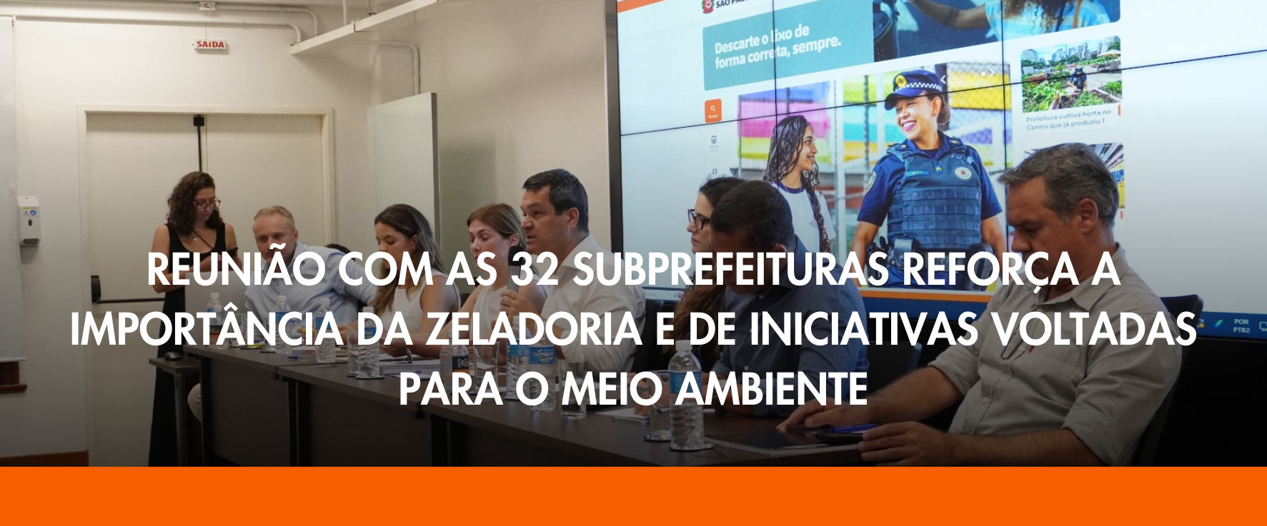 Sete pessoas estão sentadas em uma bancada. No lado esquerdo da imagem, há uma mulher branca, vestindo um vestido preto e óculos, que se encontra em pé. Ao centro da bancada, um homem branco vestindo uma camisa branca, que pode ser identificado como o Secretário Municipal Fabrício Cobra, segura um microfone em uma das mãos enquanto fala.