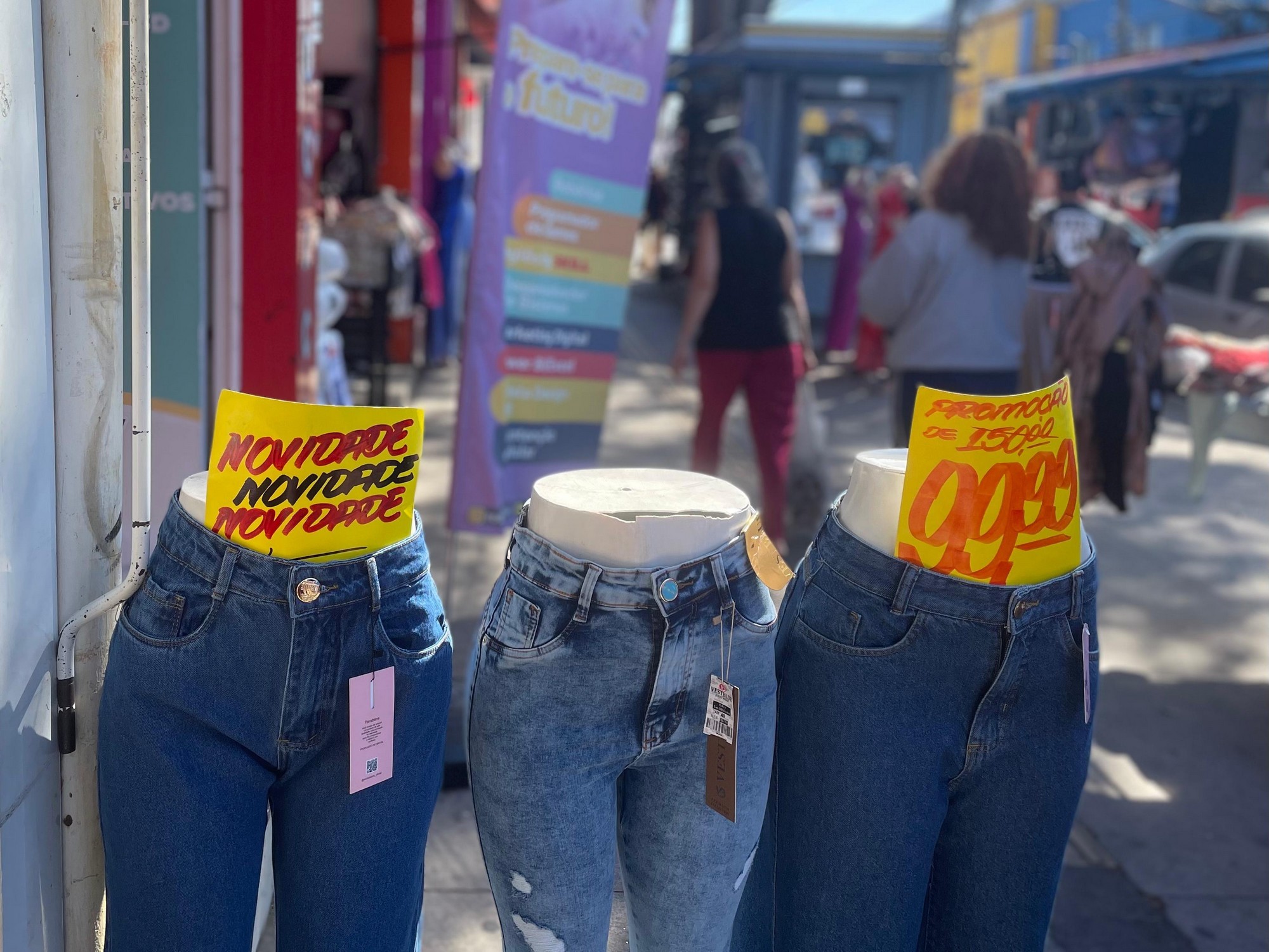 #PraTodosVerem: Três manequins vestindo calças jeans nas cores azul escuro, azul médio e azul escuro respectivamente. No primeiro, uma placa em amarelo escrito “novidade” três vezes; no terceiro, uma placa amarela indicando o valor de R$ 99,99. Ao fundo, pessoas transitando.
