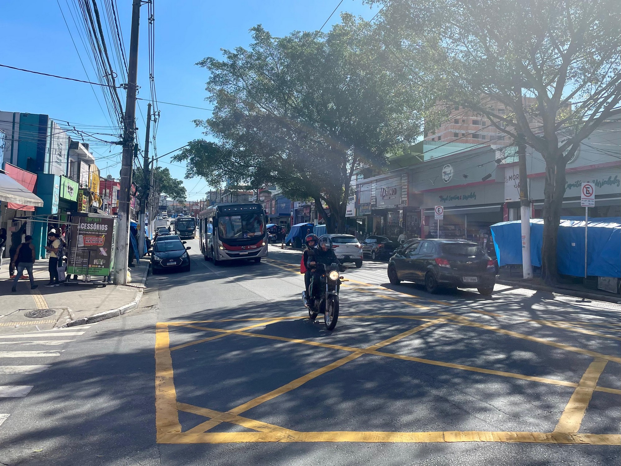 #PraTodosVerem: Imagem da esquina de uma rua, com uma faixa de pedestres aparecendo no canto inferior esquerdo. Árvores na calçada da direita, bem como barracas azuis de vendas. No centro da rua, um ônibus e uma moto.