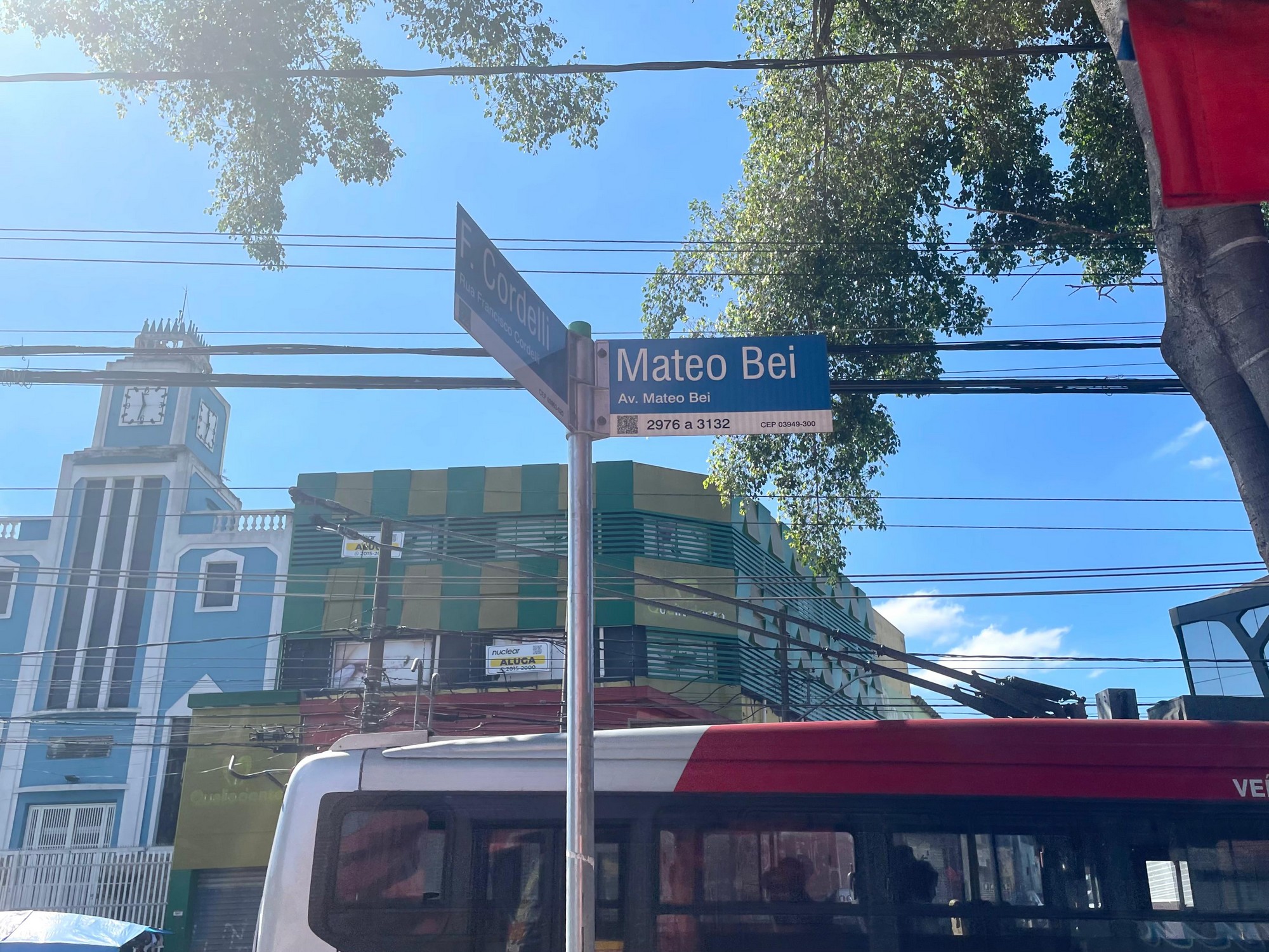 #PraTodosVerem: Ao fundo, céu azul com raios de sol iluminando a foto. Árvores aparecendo no canto superior direito da tela e, em foco na imagem, uma placa indicando a Rua Francisco Cordelli à esquerda e a Avenida Mateo Bei à direita.