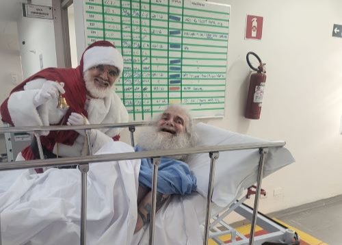 Na imagem está um homem vestido de papai noel com um sininho na mão. Ele está atrás de uma maca na qual está deitado um homem idoso com uma grande barba branca. Os dois estão sorrindo.