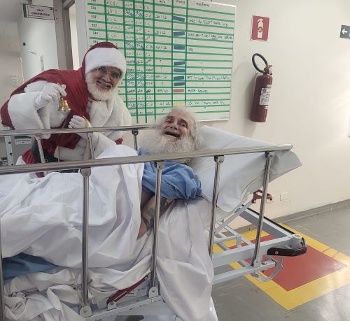 Na imagem está um homem vestido de papai noel com um sininho na mão. Ele está atrás de uma maca na qual está deitado um homem idoso com uma grande barba branca. Os dois estão sorrindo.