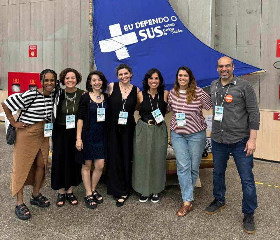 A imagem mostra um grupo de sete pessoas posando para a foto em um evento. Fernanda está à esquerda. Todos estão sorrindo e usando crachás pendurados no pescoço, indicando que participam da ocasião. Atrás do grupo, há um banner azul com a frase "EU DEFENDO O SUS - Sistema Único de Saúde", reforçando o contexto de apoio à saúde pública. O grupo é diverso em termos de vestimenta e estilo, transmitindo um clima descontraído e engajado.