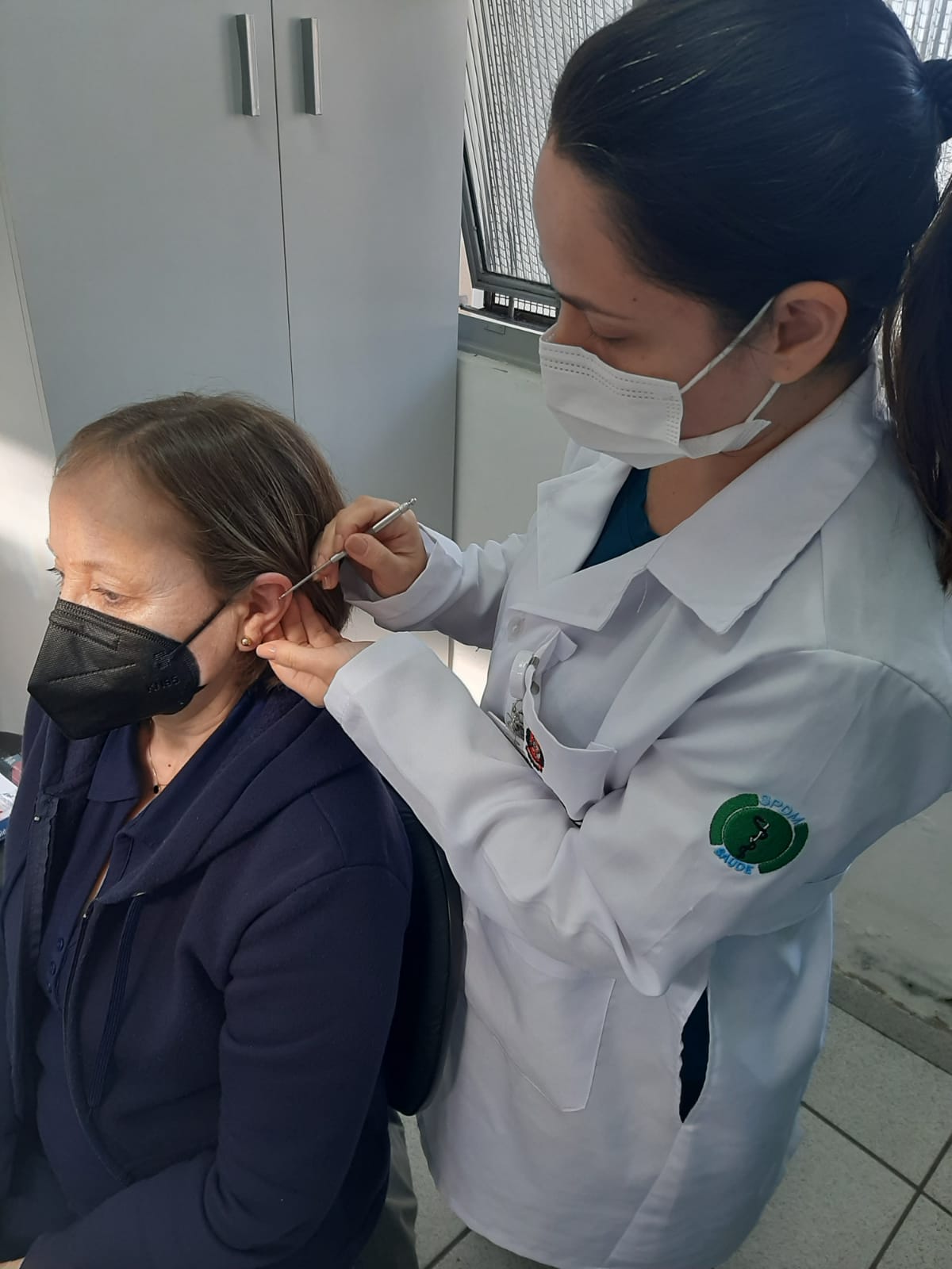 A foto mostra duas mulheres; uma está sentada, e a segunda de pé, aplicando uma espécie de agulha na orelha da outra; ambas usam máscaras 