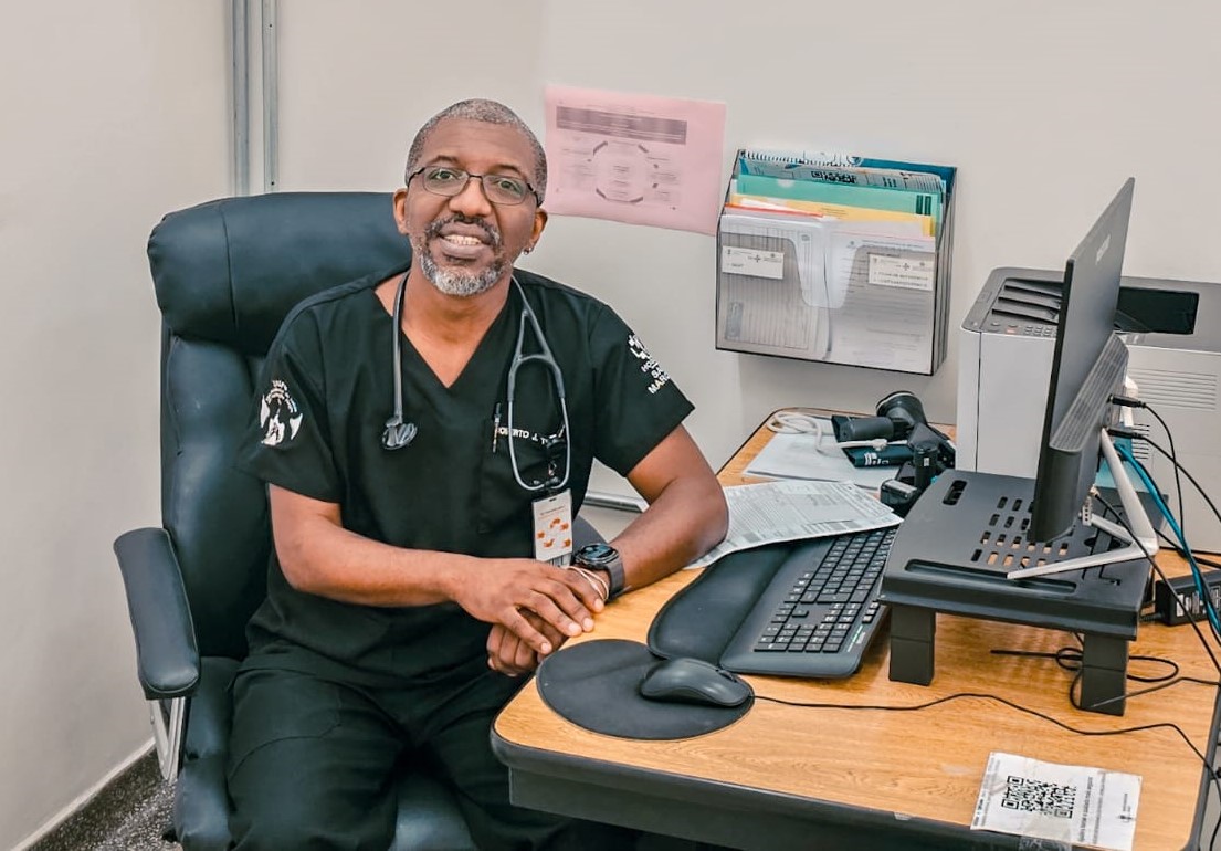  A imagem mostra o médico Roberto, um homem negro de cabelo e barba grisalhos. Ele está sentado em uma cadeira em frente a uma mesa de escritório e usa um uniforme preto com um estetoscópio pendurado no pescoço e um crachá visível. Ele está num consultório com computador, impressora, papéis organizados em suportes de parede e materiais administrativos. 