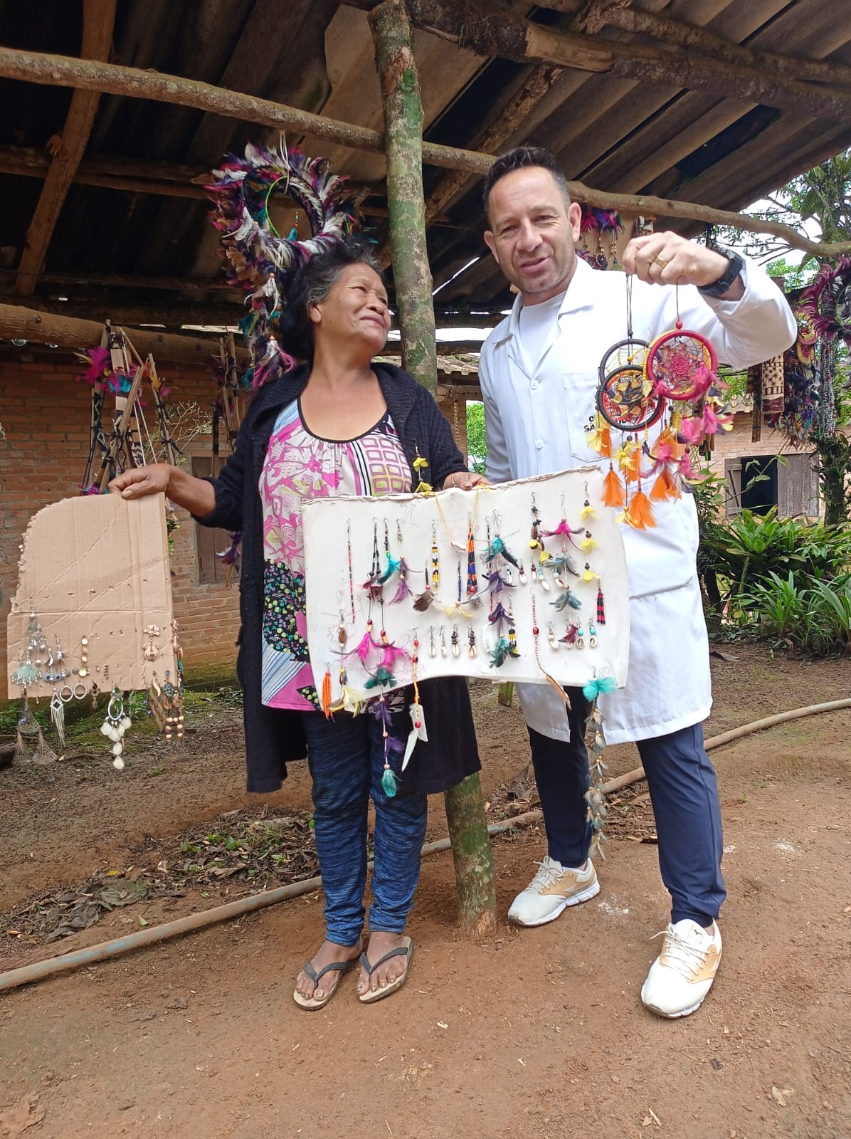 A imagem mostra duas pessoas sorrindo enquanto seguram artesanatos. À esquerda, uma mulher com cabelos pretos e longos exibe dois suportes feitos de papelão contendo brincos e outros acessórios coloridos, muitos decorados com penas. À direita, está Américo. Ele usa jaleco branco e calças escuras segura dois filtros dos sonhos coloridos e decorados com penas e fios.