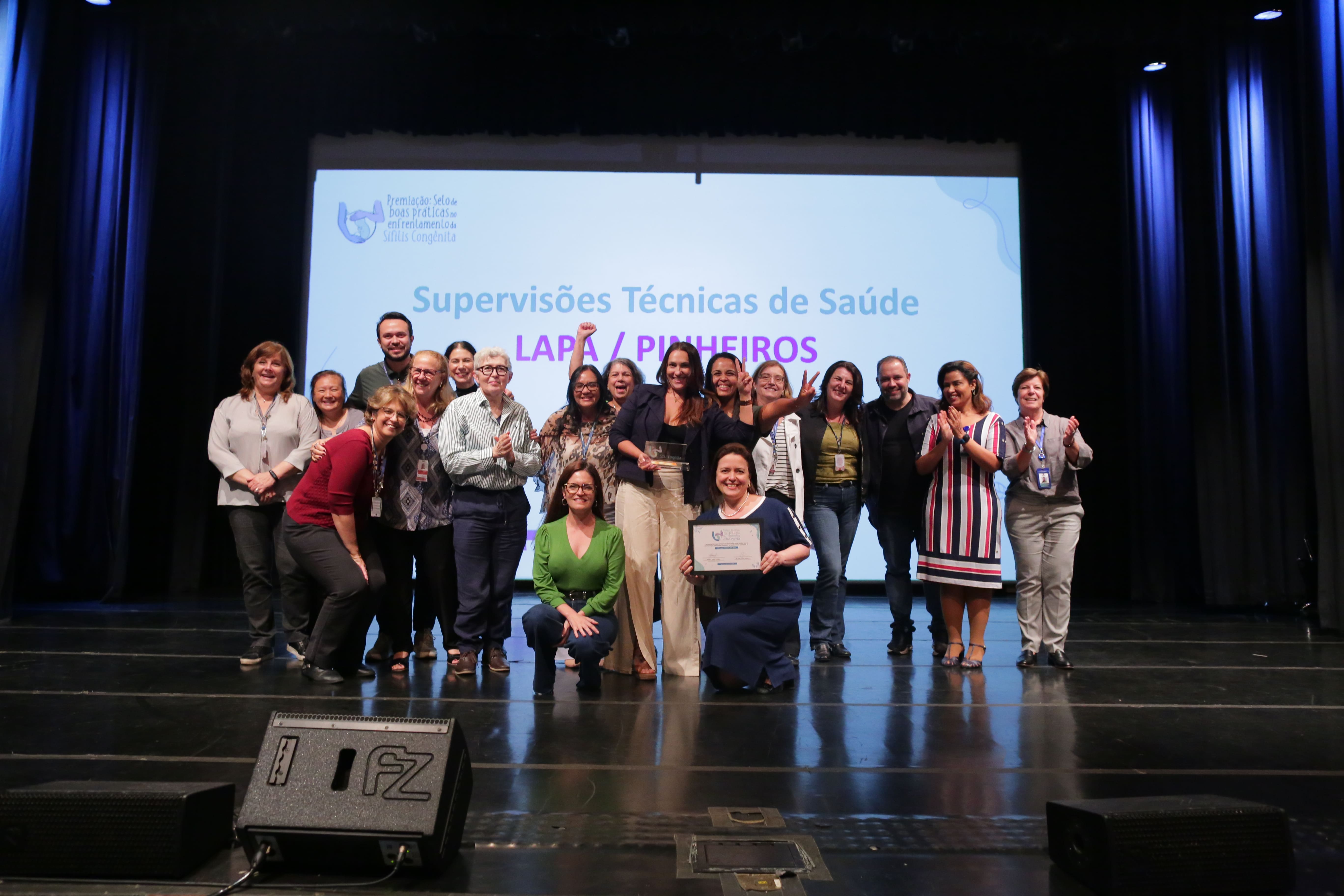 A imagem mostra um grupo de pessoas em um palco posando para a foto. Elas estão sorrindo e aparentam estar celebrando uma premiação ou evento. No fundo, há uma tela de projeção que exibe o texto: "Supervisões Técnicas de Saúde LAPA / PINHEIROS". A projeção também menciona "Premiação Selo de Boas Práticas em Enfrentamento à Sífilis Congênita". Uma das pessoas na frente segura um certificado