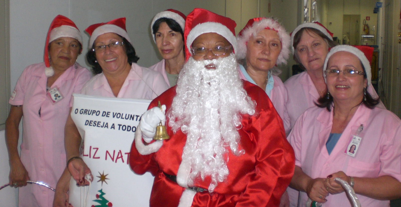  A imagem mostra um grupo de sete pessoas, sendo seis mulheres e uma delas vestida como Papai Noel no centro. As mulheres estão usando chapéus de Papai Noel e uniformes em tons de rosa claro. Uma delas segura um cartaz com a mensagem: "O GRUPO DE VOLUNTÁRIOS DESEJA A TODOS UM FELIZ NATAL." 