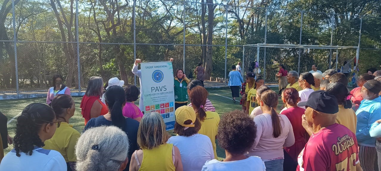A imagem mostra uma ação comunitária ao ar livre realizada em uma quadra esportiva cercada por alambrado, possivelmente em um parque. Um grupo grande de pessoas, composto majoritariamente por adultos e idosos, está reunido em torno de uma mulher que segura um banner do PAVS – Programa Ambientes Verdes e Saudáveis, da Secretaria Municipal da Saúde.  O banner traz o título “Saúde Ambiental” e menciona o combate às arboviroses, além de exibir ícones dos Objetivos de Desenvolvimento Sustentável (ODS) da ONU, sinalizando o compromisso com a saúde e a sustentabilidade. Algumas pessoas vestem camisetas com mensagens ou logotipos de grupos organizados, indicando participação ativa da comunidade.  Ao fundo, é possível ver mais pessoas caminhando ou interagindo em um espaço arborizado, reforçando o caráter coletivo, educativo e de promoção da saúde do evento. 