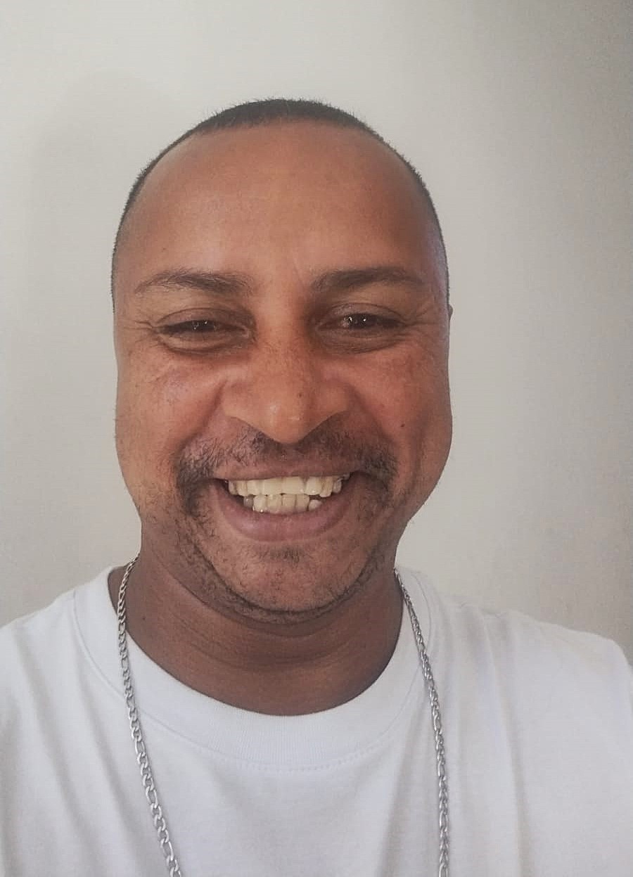 A imagem mostra um homem sorrindo amplamente em um fundo claro e neutro. Ele tem cabelo curto, usa uma camiseta branca e uma corrente prateada no pescoço. Seu sorriso expressa felicidade e seus olhos estão ligeiramente fechados, destacando o momento descontraído e alegre. 